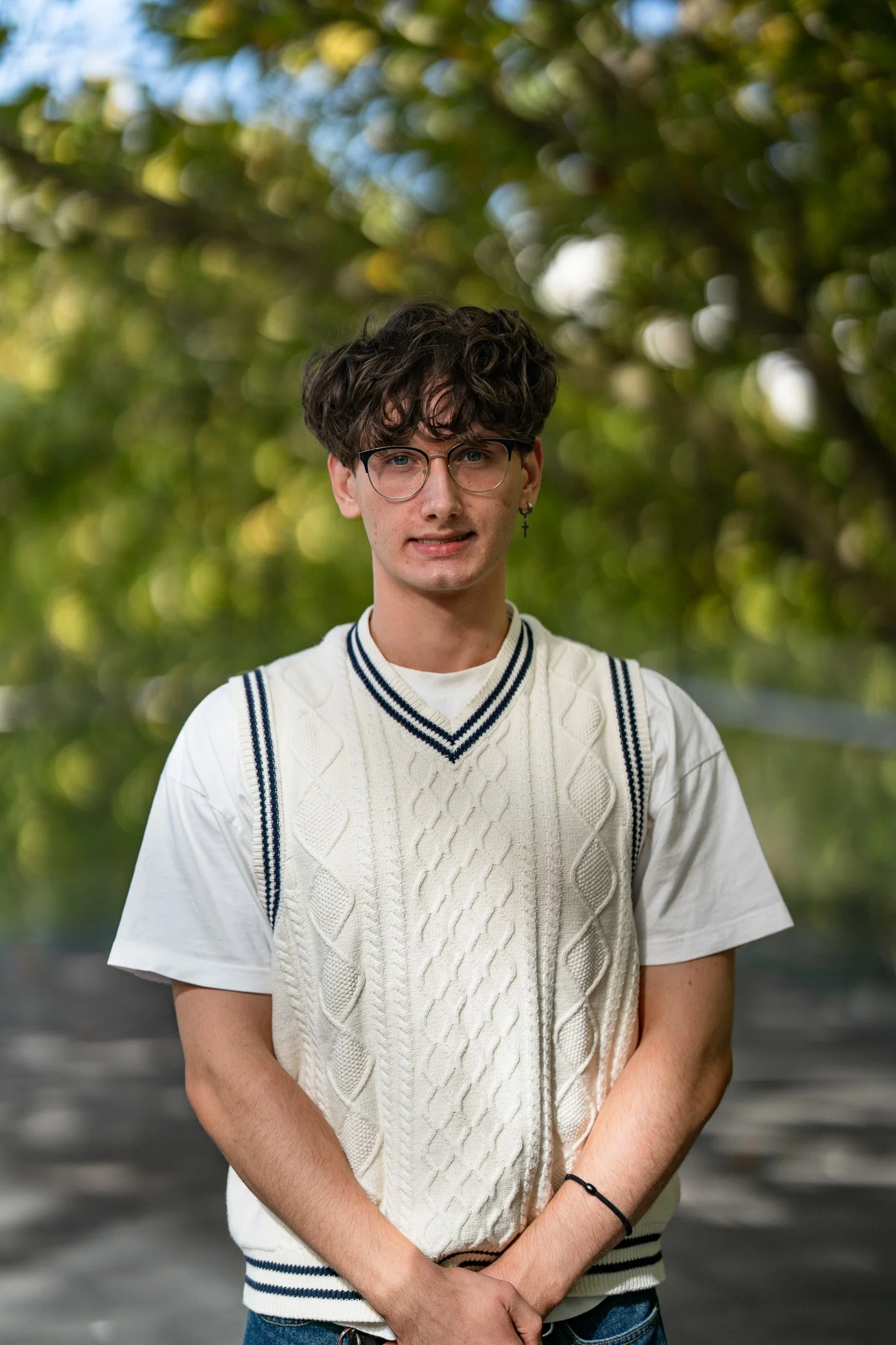 A young man with curly dark hair, glasses, and a cross earring, standing outdoors with blurred green foliage in the background, wearing a white short-sleeved shirt and a cream-colored cable-knit vest with navy blue stripes.