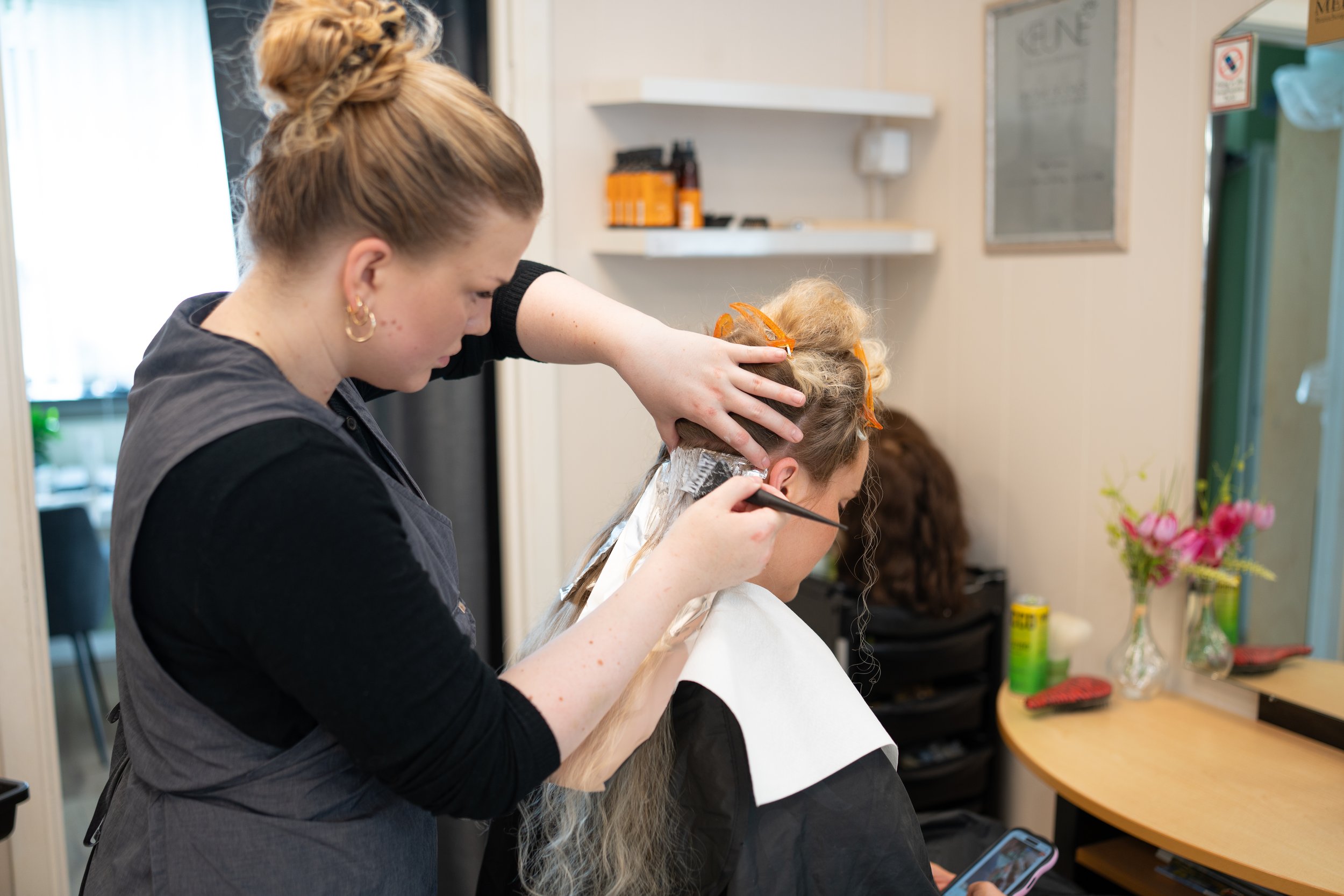 A hairstylist applying hair color to a client's hair in a salon, with the client looking at her phone.