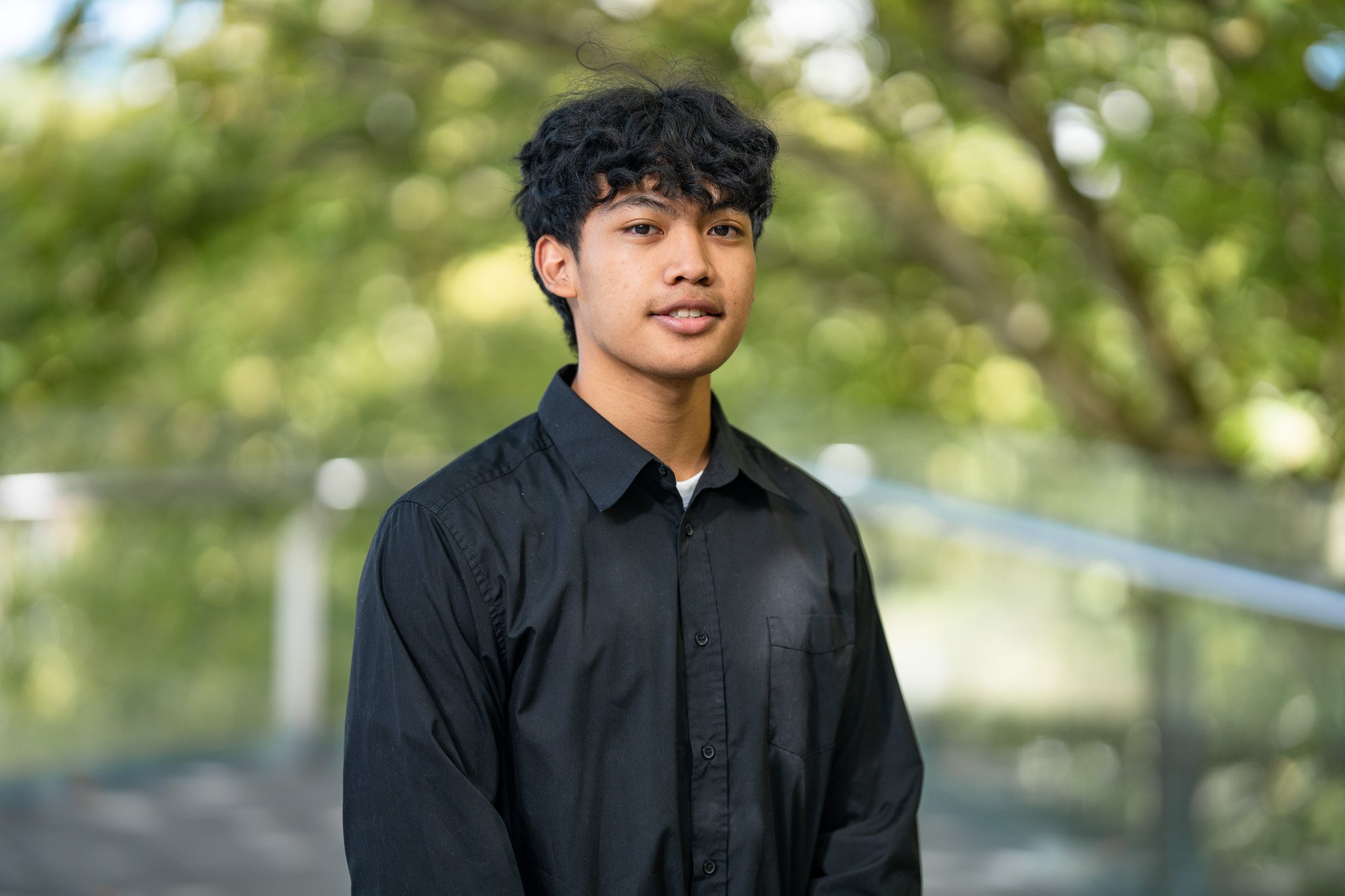 A young man with dark curly hair, wearing a black button-up shirt, standing outdoors with green trees and a blurred background.