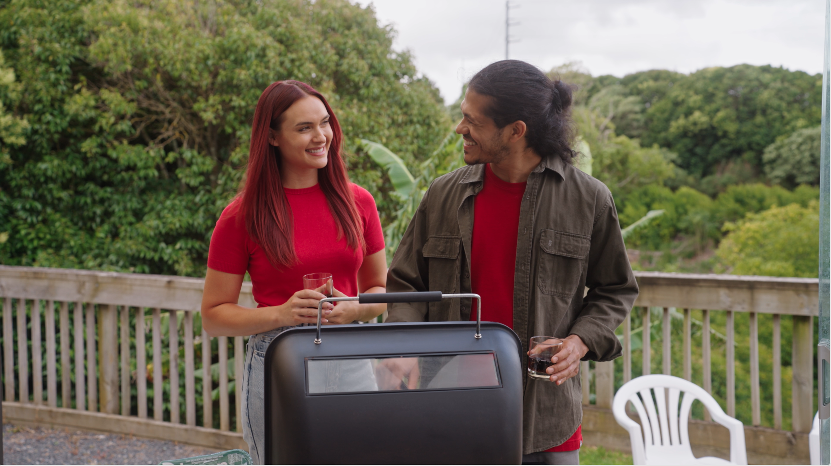 Two people, a woman with red hair and a man with long dark hair, are smiling and talking outdoors near a grill, holding glasses of drinks, with green trees and a wooden fence in the background.