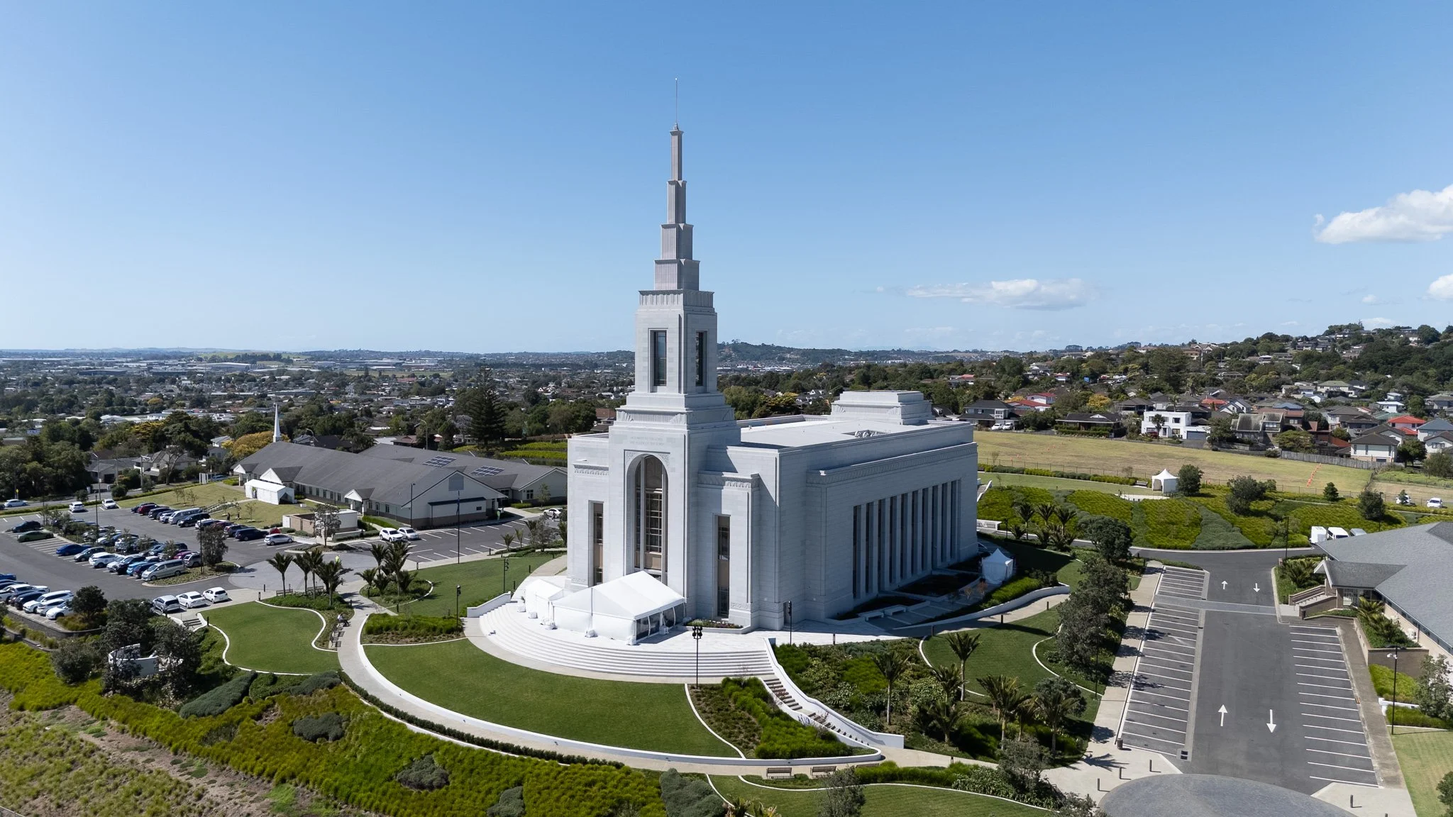 A white church with a tall steeple on a hilltop, surrounded by parking lots and green landscaping, with a clear blue sky and a sprawling neighborhood in the background.