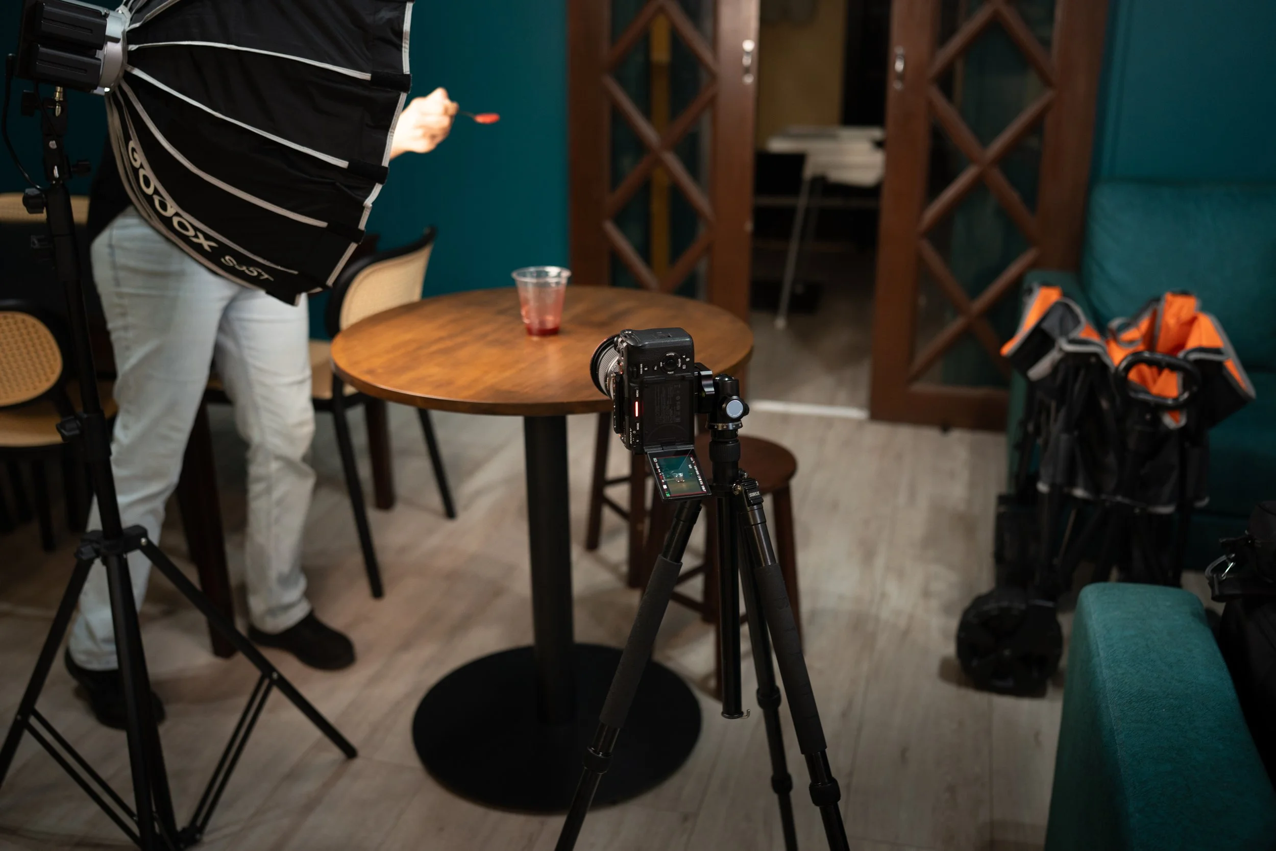 Photograph of a photography setup inside a restaurant, with a camera on a tripod focused on a table with a drink, and a person holding a reflector and a spoon.