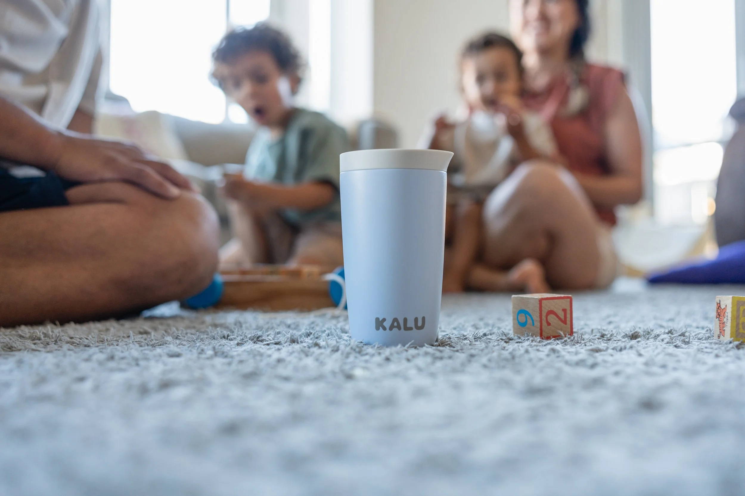 A family playing a game on the carpeted floor with game blocks and a white cup labeled "KALU" in the center, with children and a woman in the background.