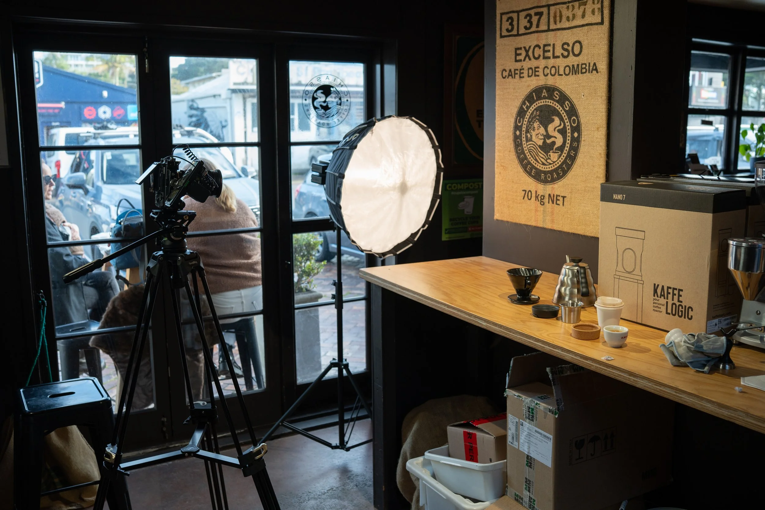 Interior view of a coffee shop with a camera and lighting setup for filming, a wooden countertop with coffee brewing equipment, boxes, and cafe packaging, large windows showing parked cars outside.