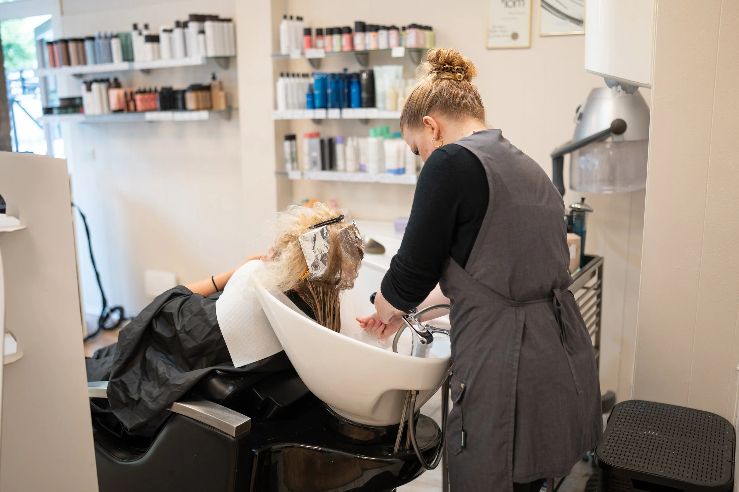 A hairdresser washing a young girl's hair at a salon sink while the girl leans back with a hair cap and towel on her head, surrounded by shelves of hair products.
