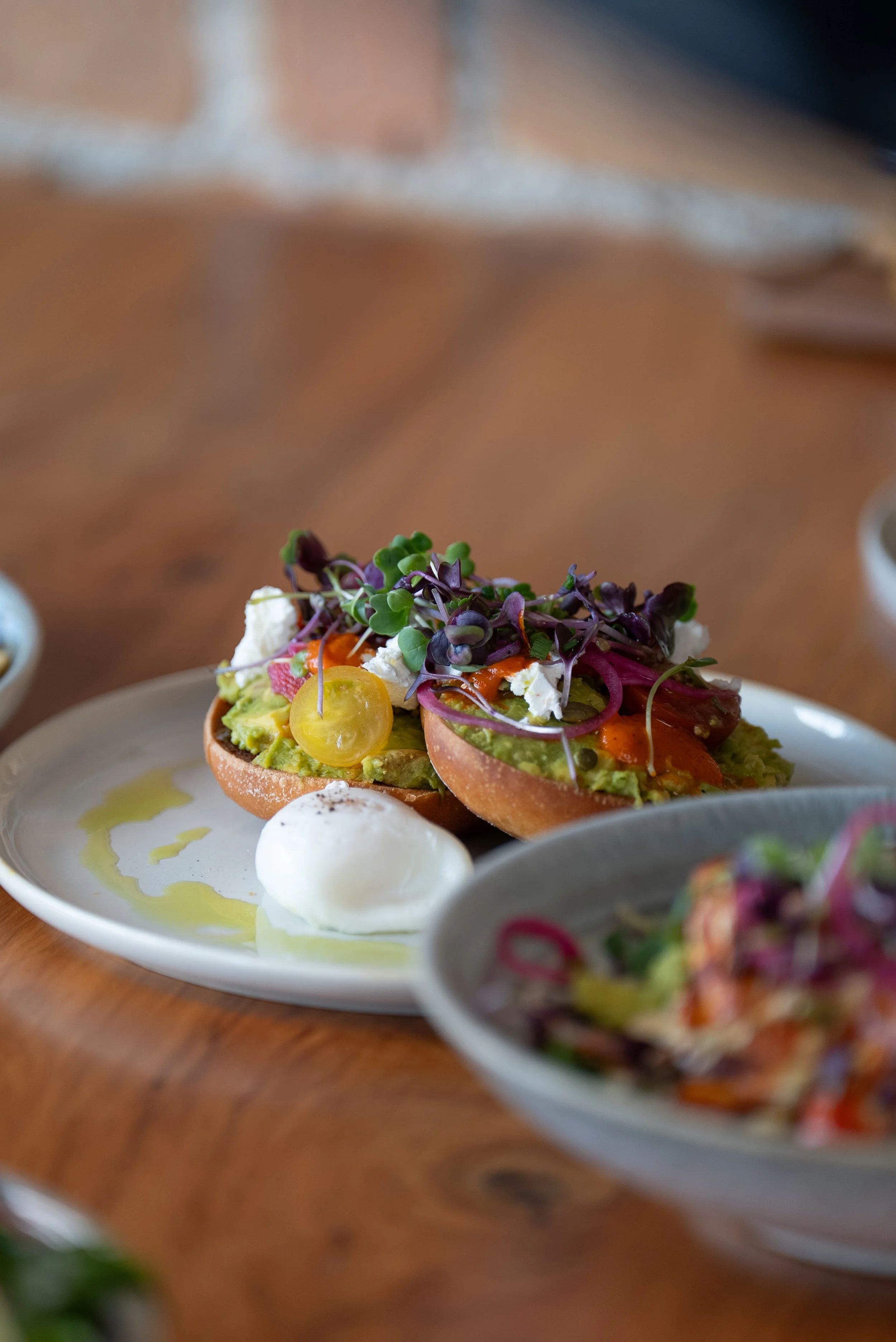 Close-up of avocado toast with microgreens, cherry tomato, and feta cheese on a white plate, with a soft-boiled egg nearby, on a wooden table.