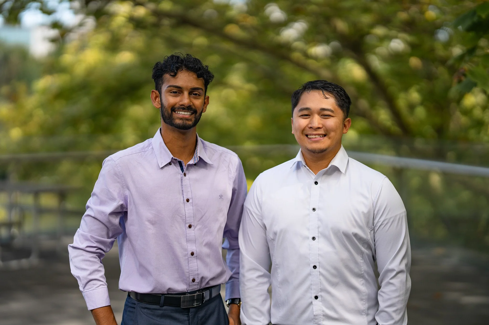 Two young men standing outside in front of trees, smiling at the camera, wearing collared shirts, one in light purple and the other in white.