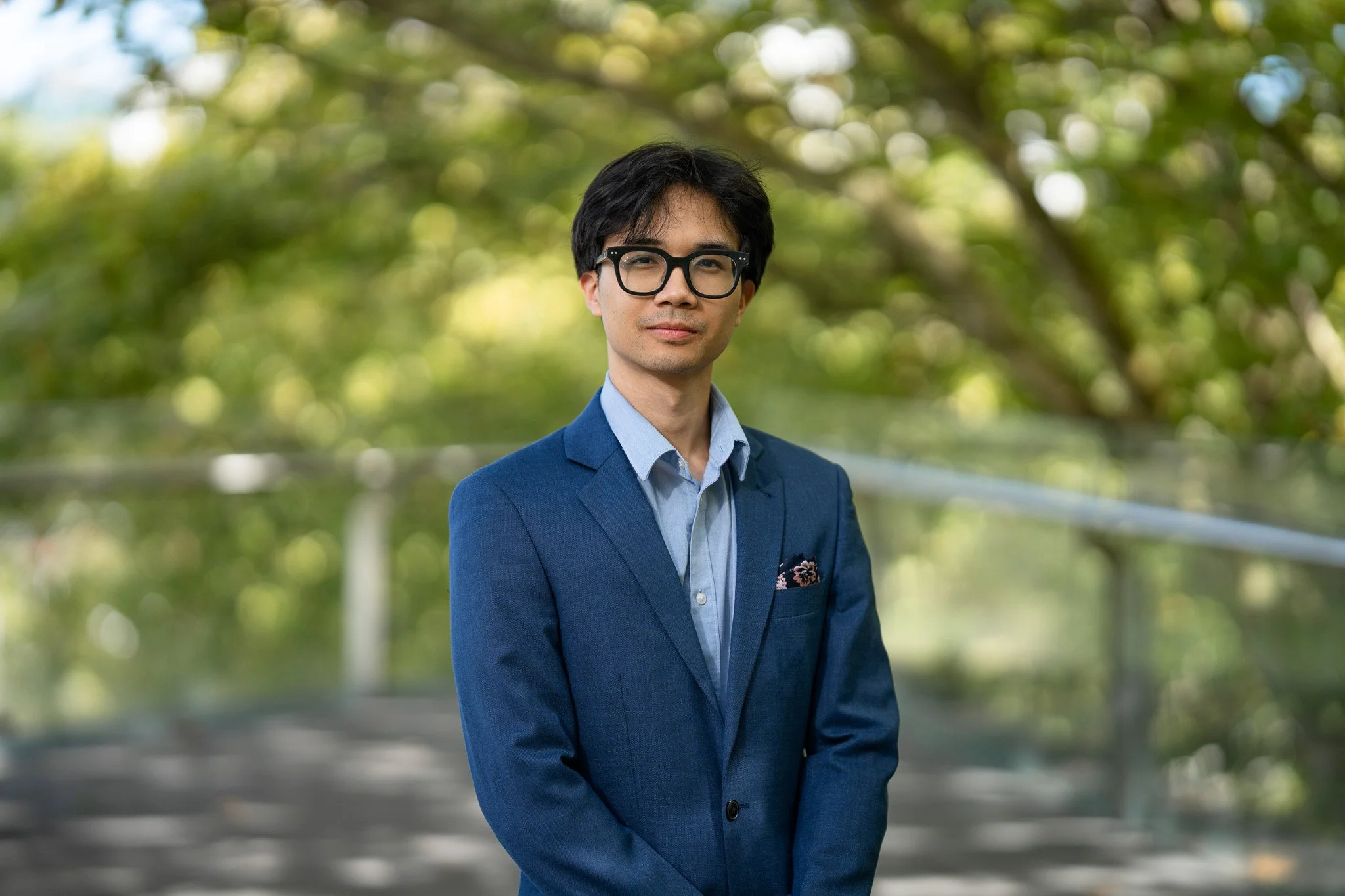 A young man with black hair, wearing glasses, a blue suit, and a light blue shirt, standing outdoors with greenery in the background.