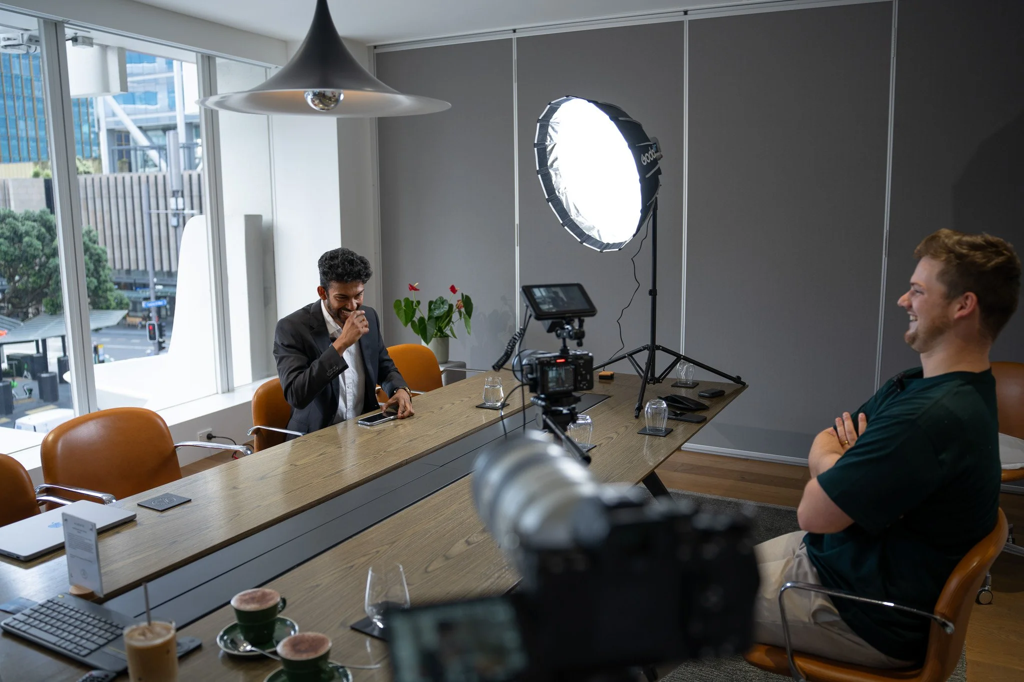 A video interview being recorded in a modern office conference room with two men, one in a suit and one in a casual shirt, sitting at a wooden table. The man in the suit is laughing while the other man is smiling with arms crossed. Equipment includes