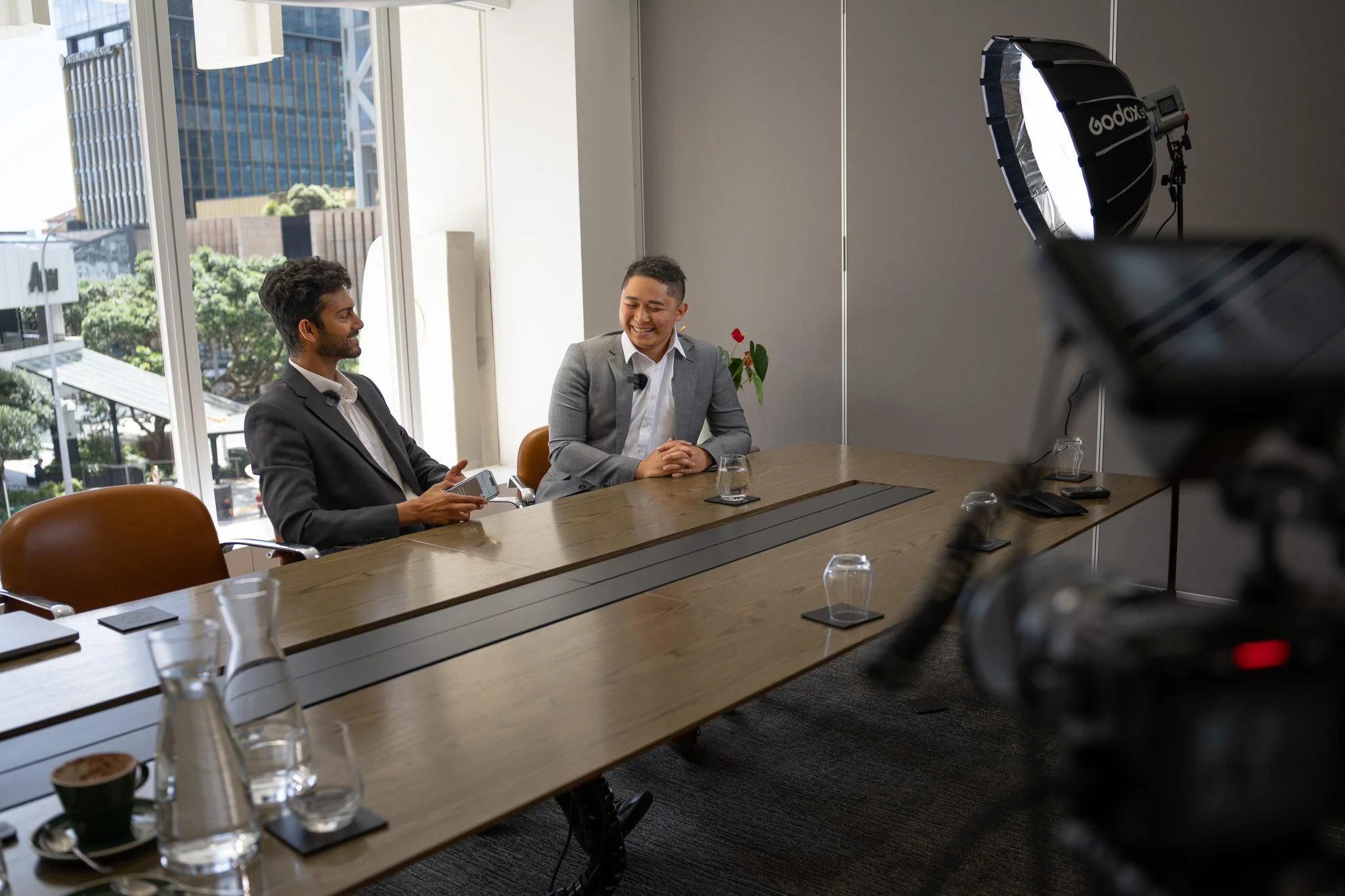 Two men in suits sitting at a conference table during an interview or discussion in a modern office with large windows and city view.