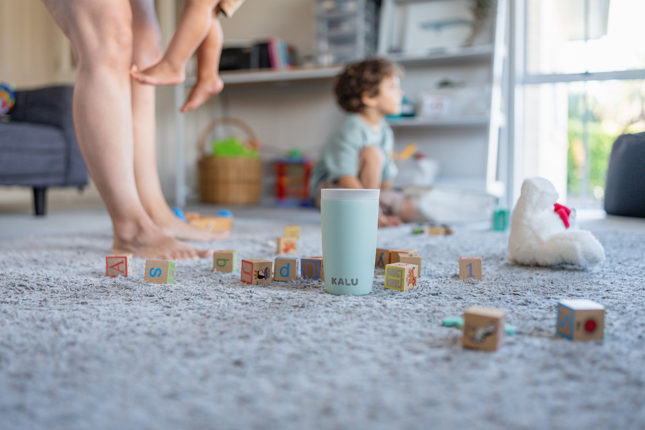 Children playing on a carpeted room with toys, including alphabet blocks and a white plush toy, while one child stands and another child sits near a window.
