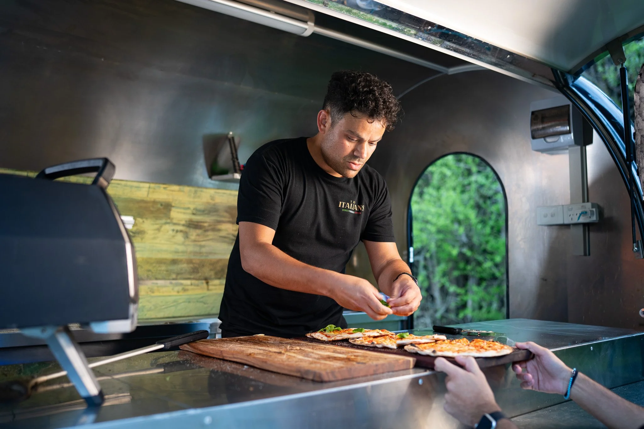 A man preparing pizza inside a mobile food truck, with an outdoor view of trees through the truck window.