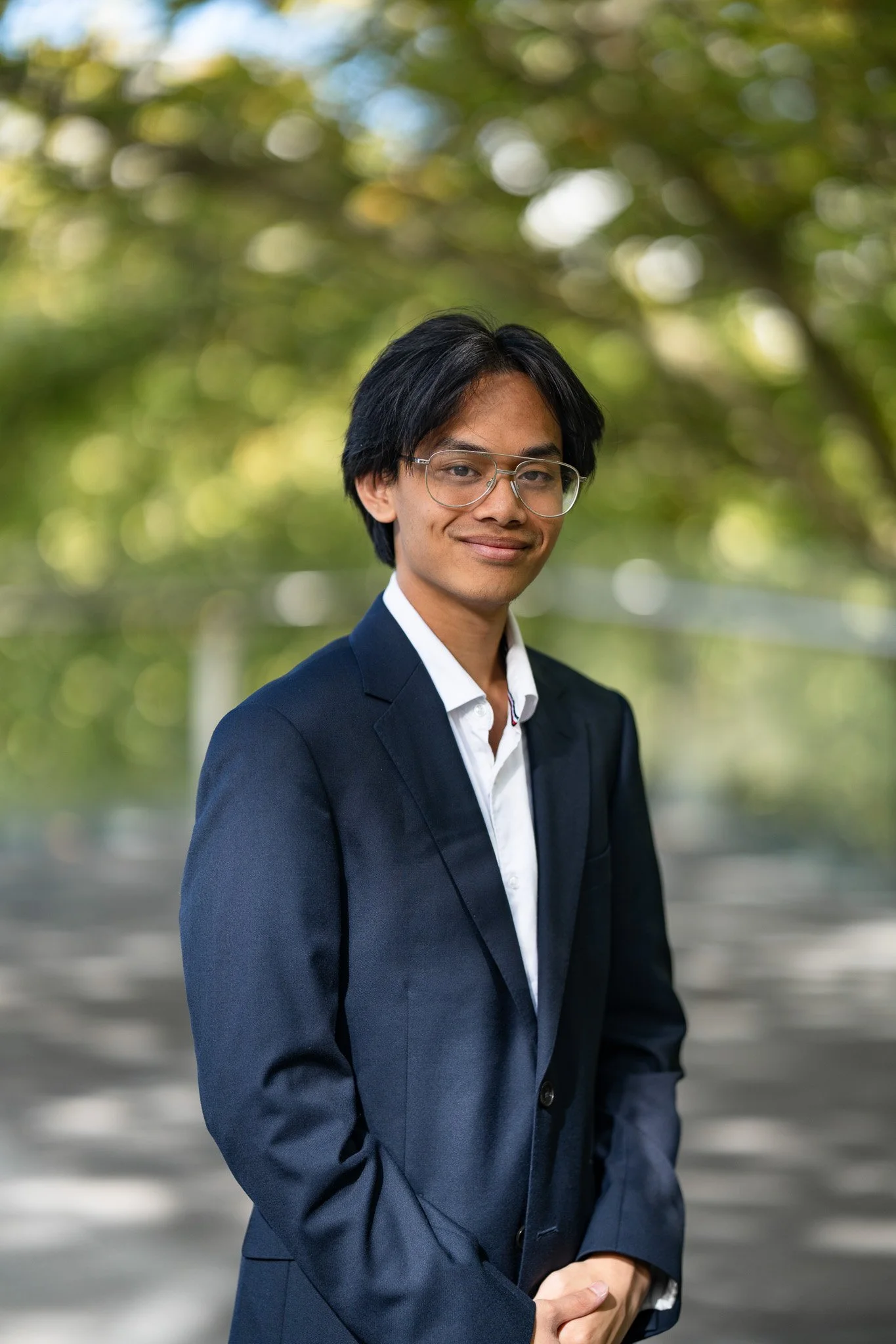 A young man with glasses wearing a dark navy suit and white shirt, standing outdoors with trees and blurred greenery in the background.