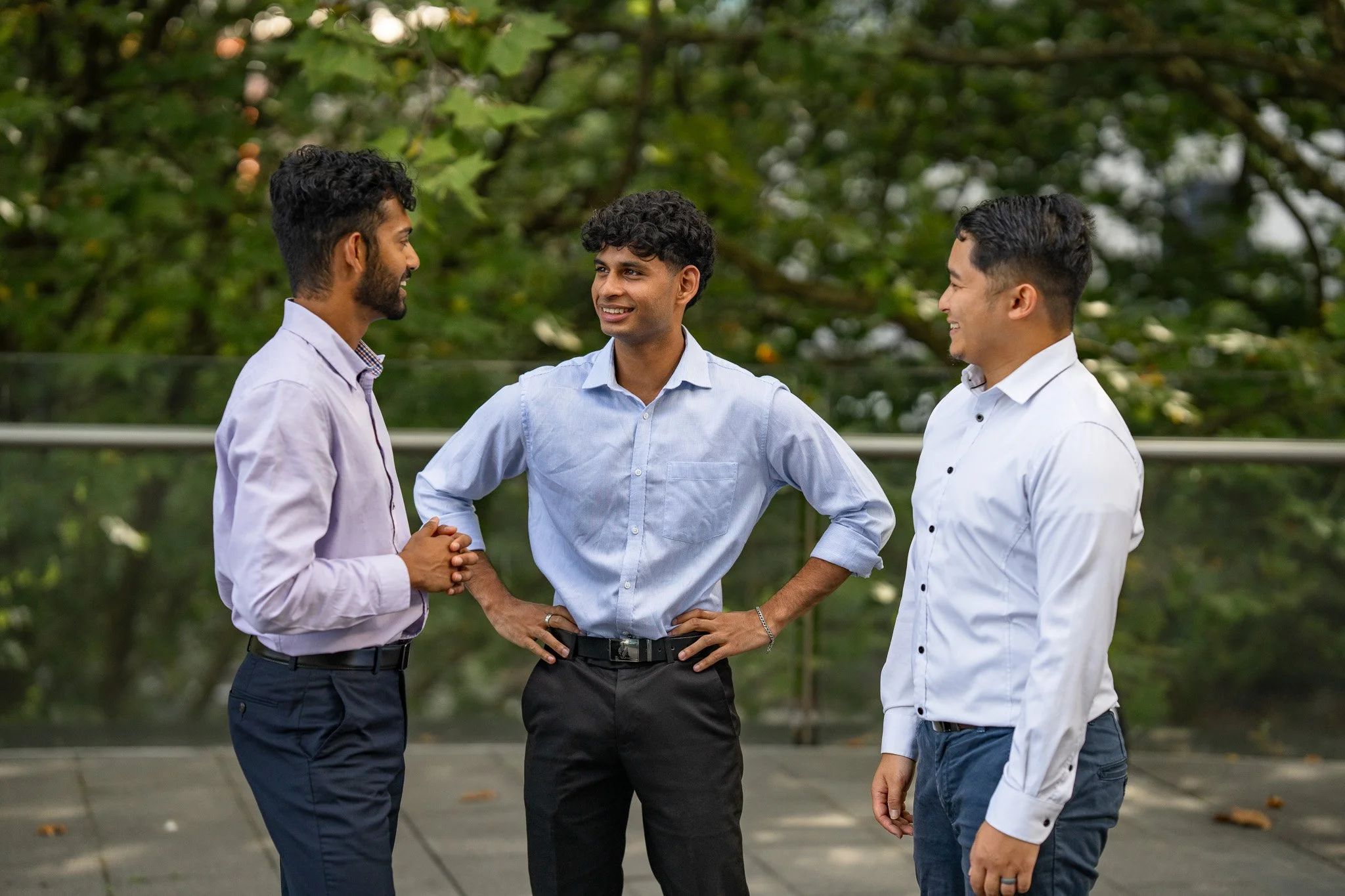 Three men talking outside on a terrace with trees in the background, all dressed in business casual attire, smiling and engaging in conversation.