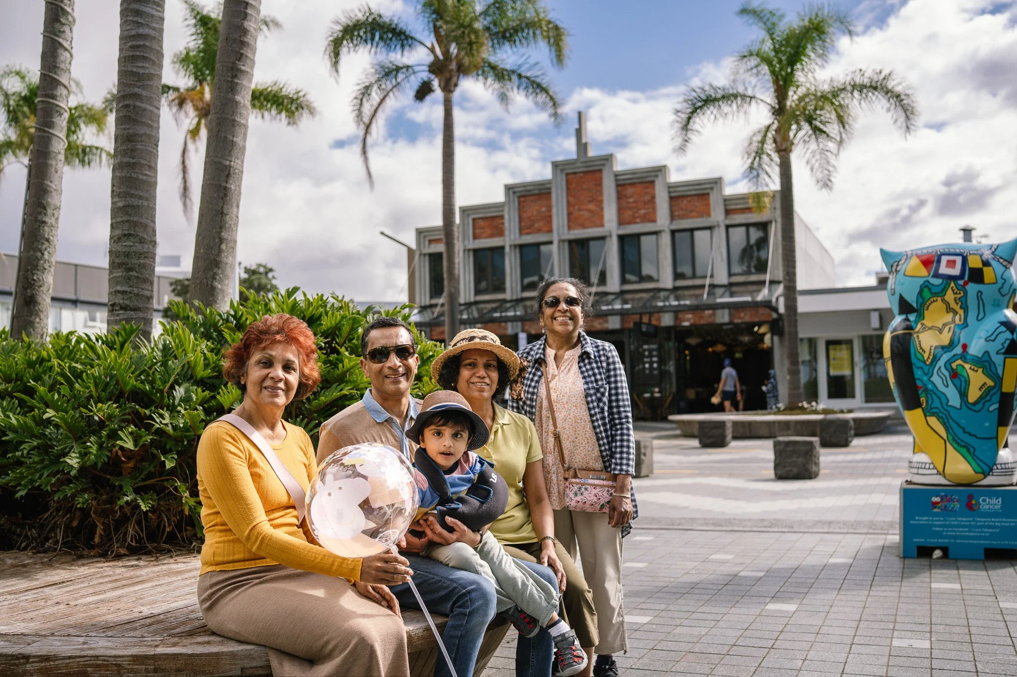 Group of five people, including a young boy, posing for a photo outdoors on a bench in front of palm trees and a building, with a sculpture in the background.
