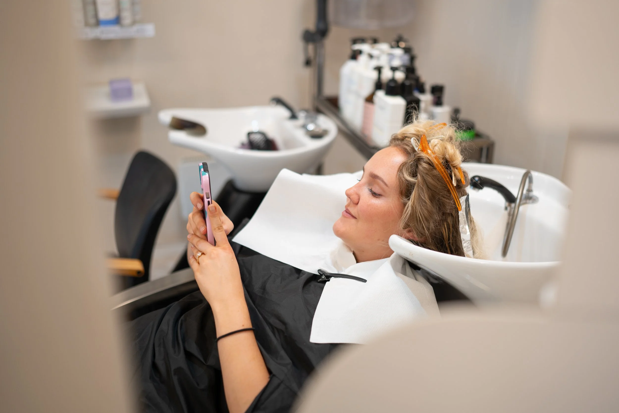 Woman with blonde hair relaxing at a salon, lying back in a salon chair with her hair covered in curlers and shades, holding a cellphone, with hair wash stations and shelves of hair products visible in the background.