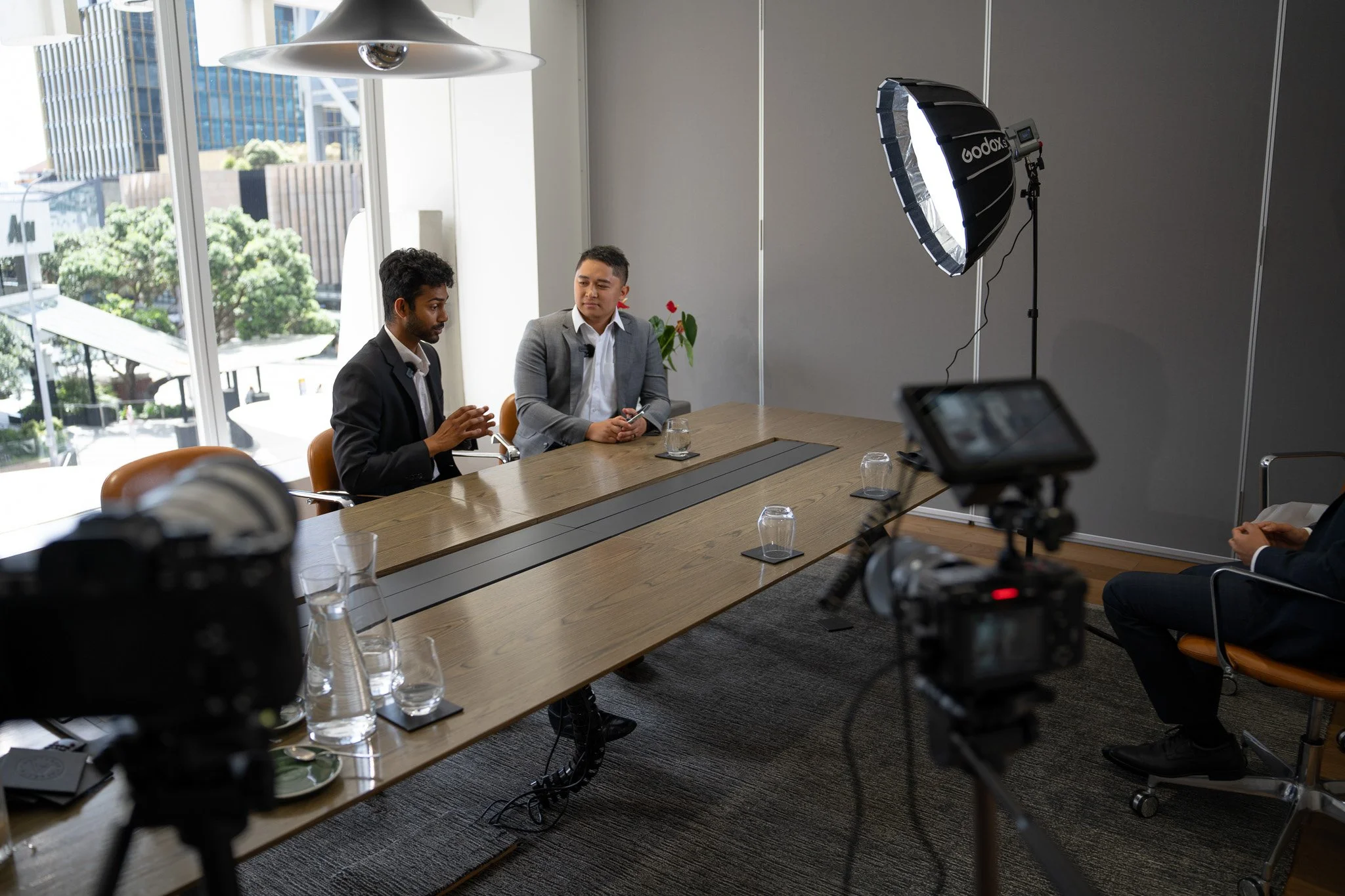 Two men sit at a conference table during an interview recording, with cameras, lighting equipment, and a large window showing city buildings in the background.