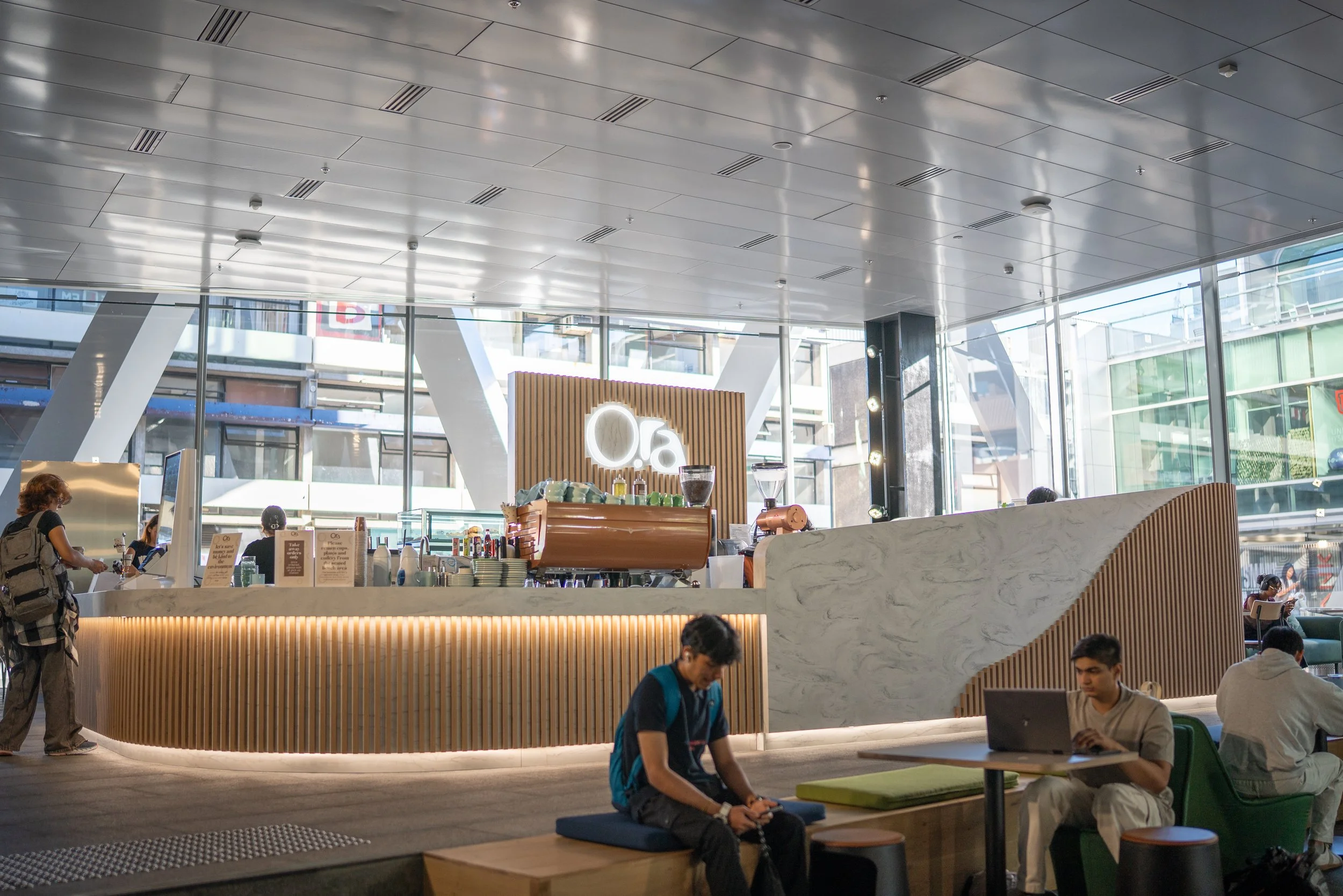 Interior of a modern coffee shop with a counter, people sitting and working, large windows, and a sign with the number '06' behind the counter.
