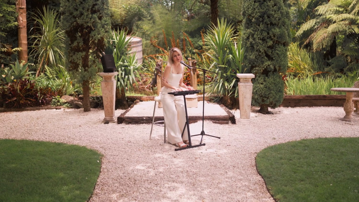 A woman in a white dress is sitting on a stool, playing a keyboard and singing into a microphone in a garden with lush green plants and trees.