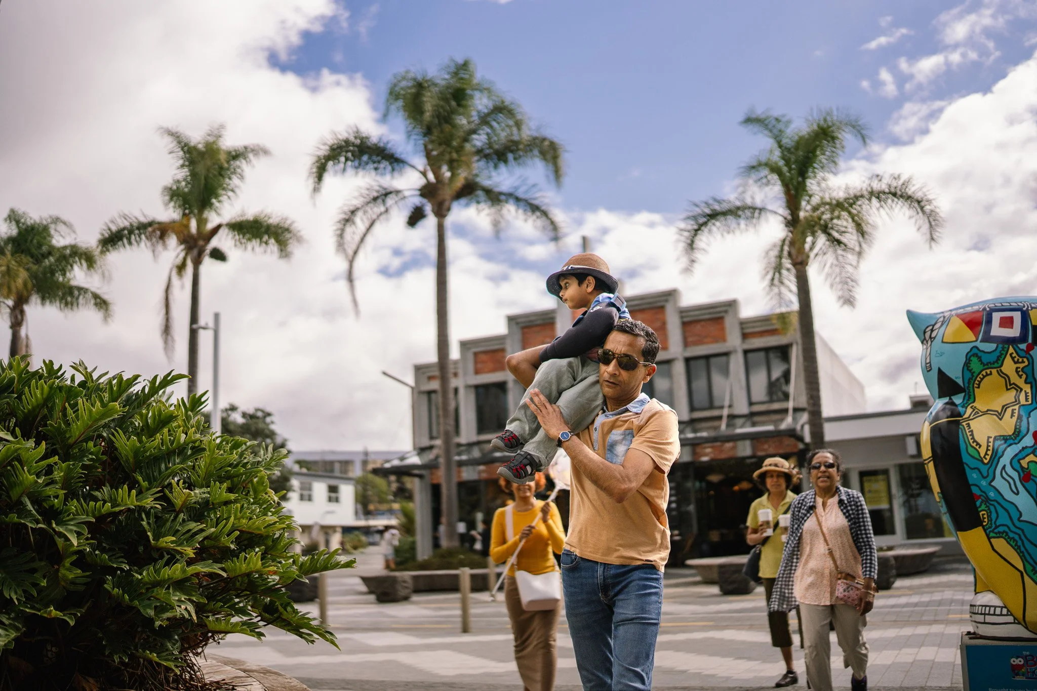 A man carrying a young boy on his shoulder walking in an outdoor shopping area with palm trees and a modern building in the background.
