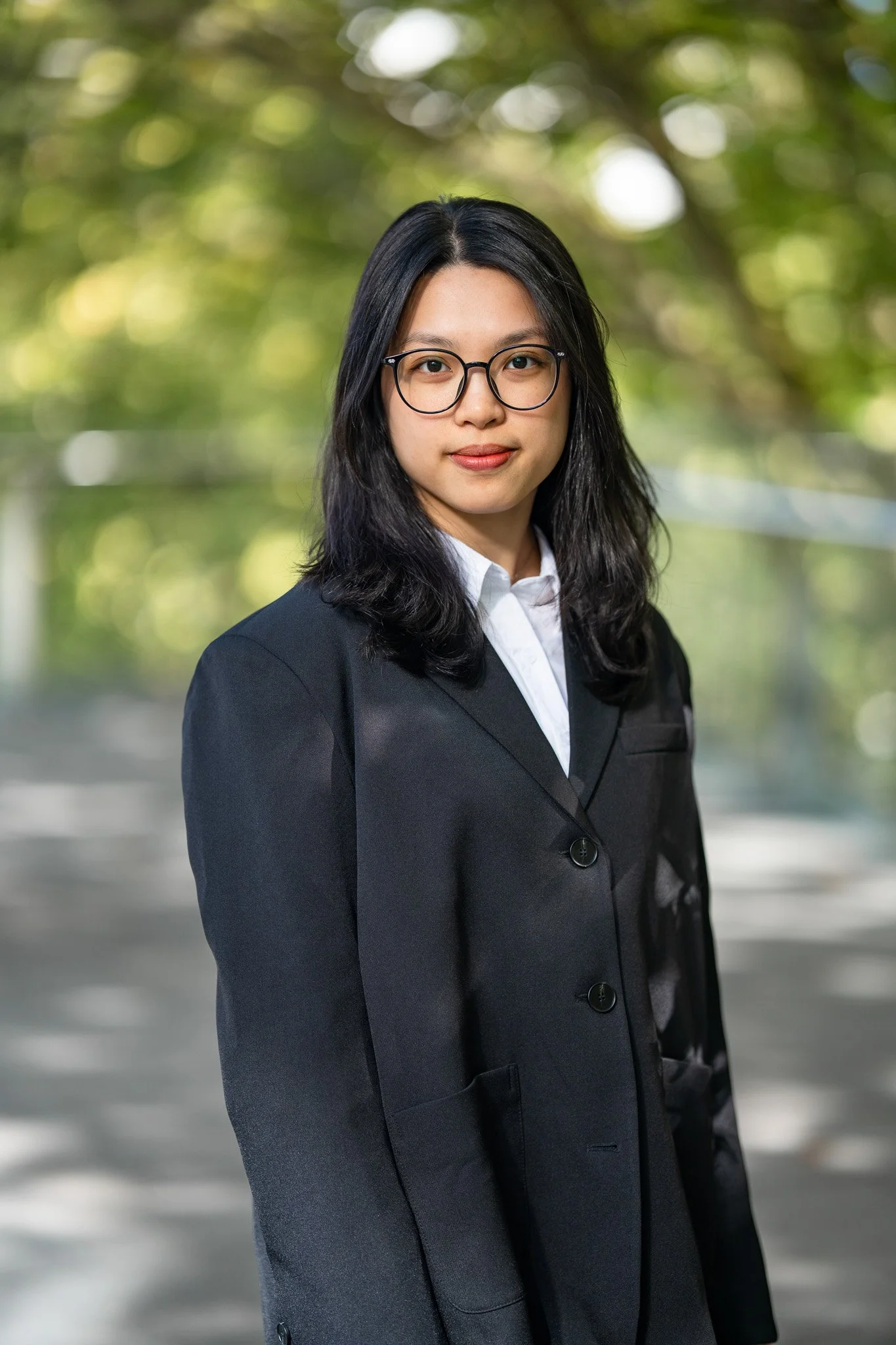 A young woman with shoulder-length black hair, wearing glasses, a black blazer, and a white shirt, standing outdoors with a blurred background of trees and sunlight.