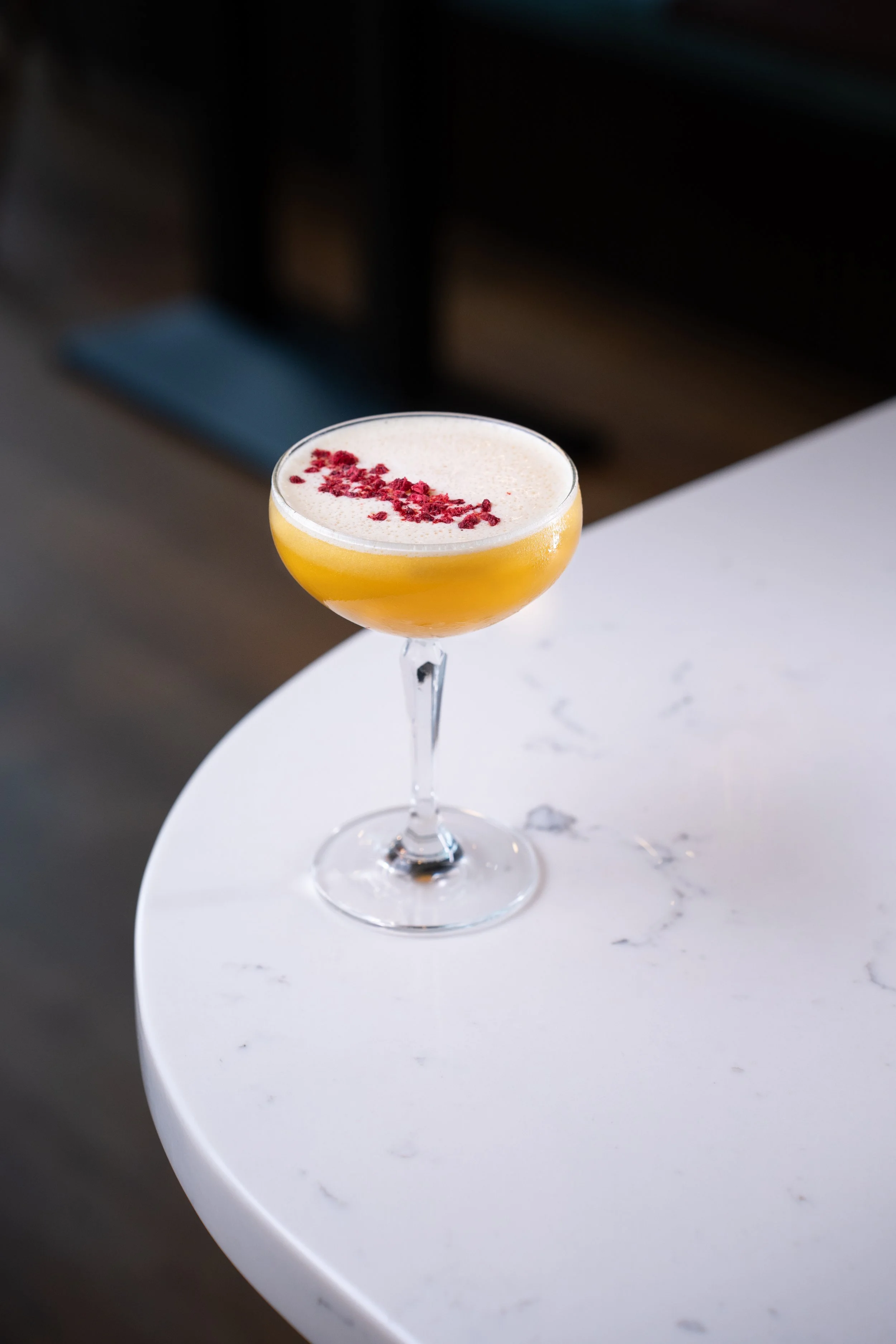 A cocktail with a frothy top and red garnish in a coupe glass on a white marble table.