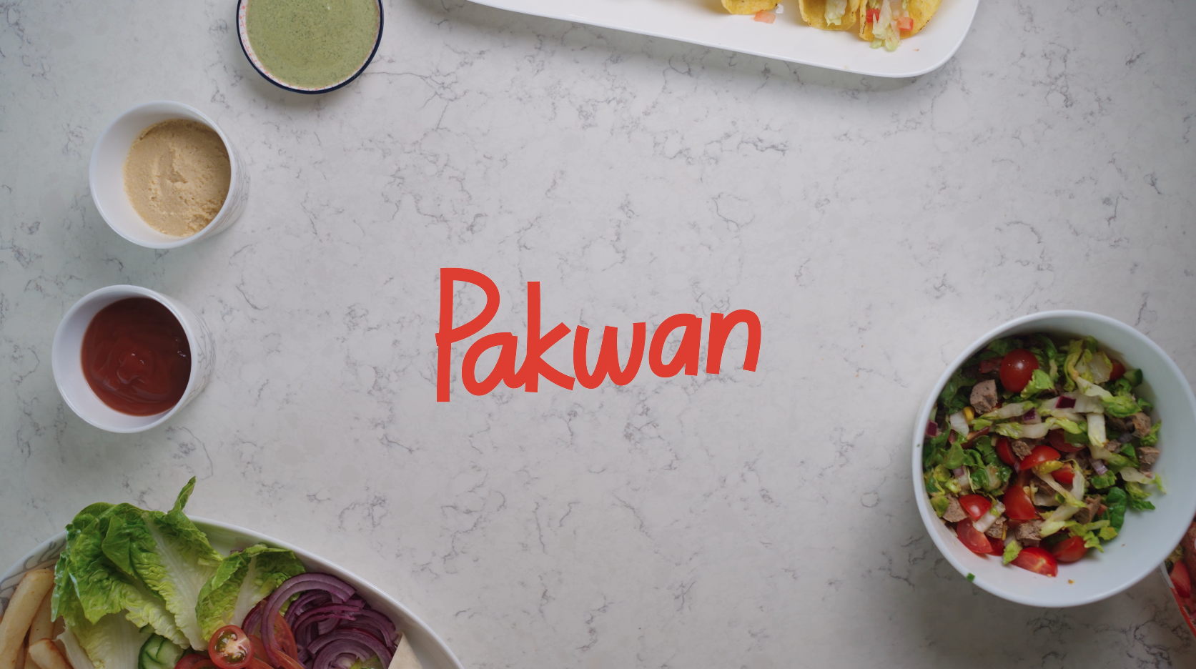 A flat lay of salads, dressings, and sauces on a white marble surface with the word 'Pakwan' in the center.