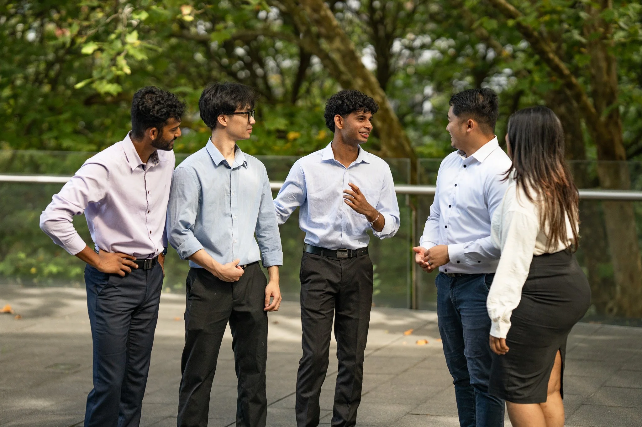 Five diverse people conversing outdoors on a paved area with trees in the background, engaged in a friendly discussion.