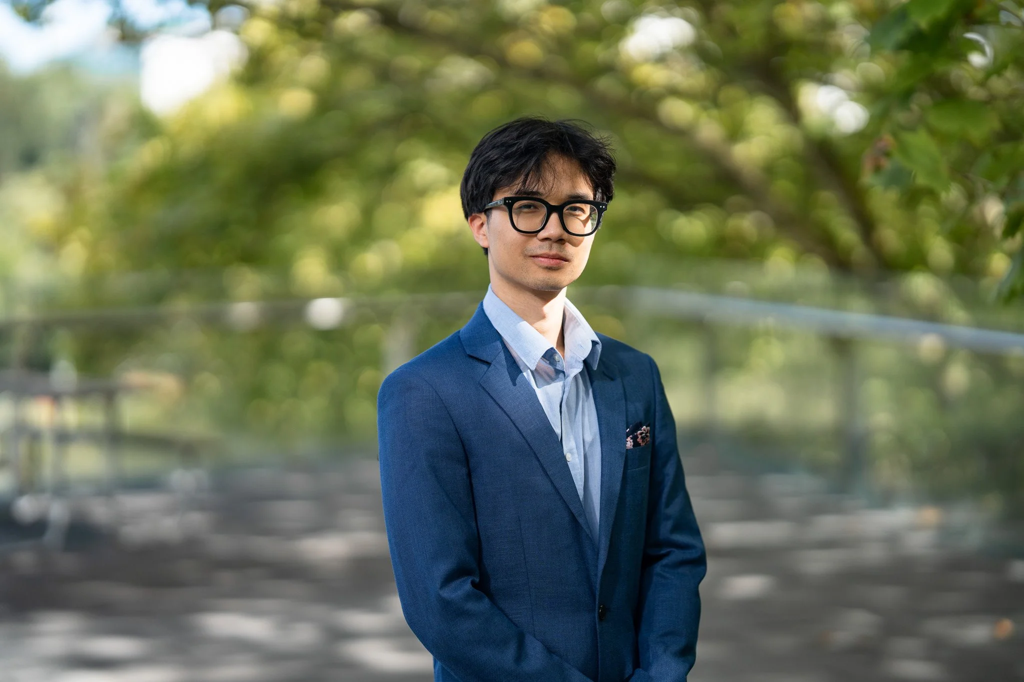 A young man dressed in a blue suit and light blue shirt standing outdoors with trees and greenery in the background.