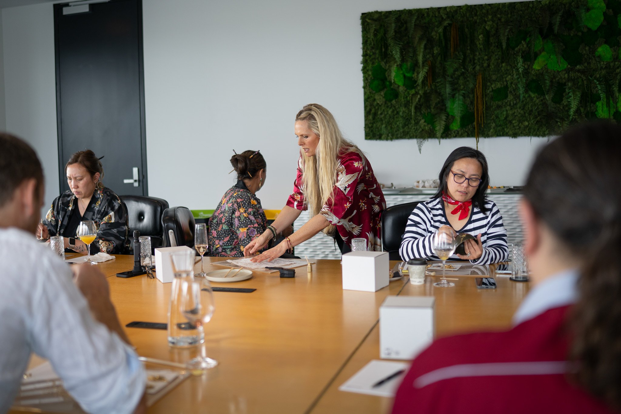 A woman in a red floral blouse teaching a table of women about sushi, with some women taking notes and wearing casual attire, in a room with a green plant wall.