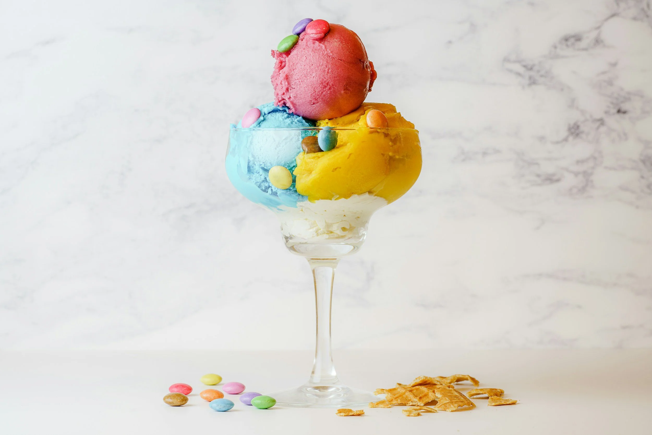 A tall glass containing three scoops of colorful ice cream, topped with small candy-coated chocolates, against a white background.