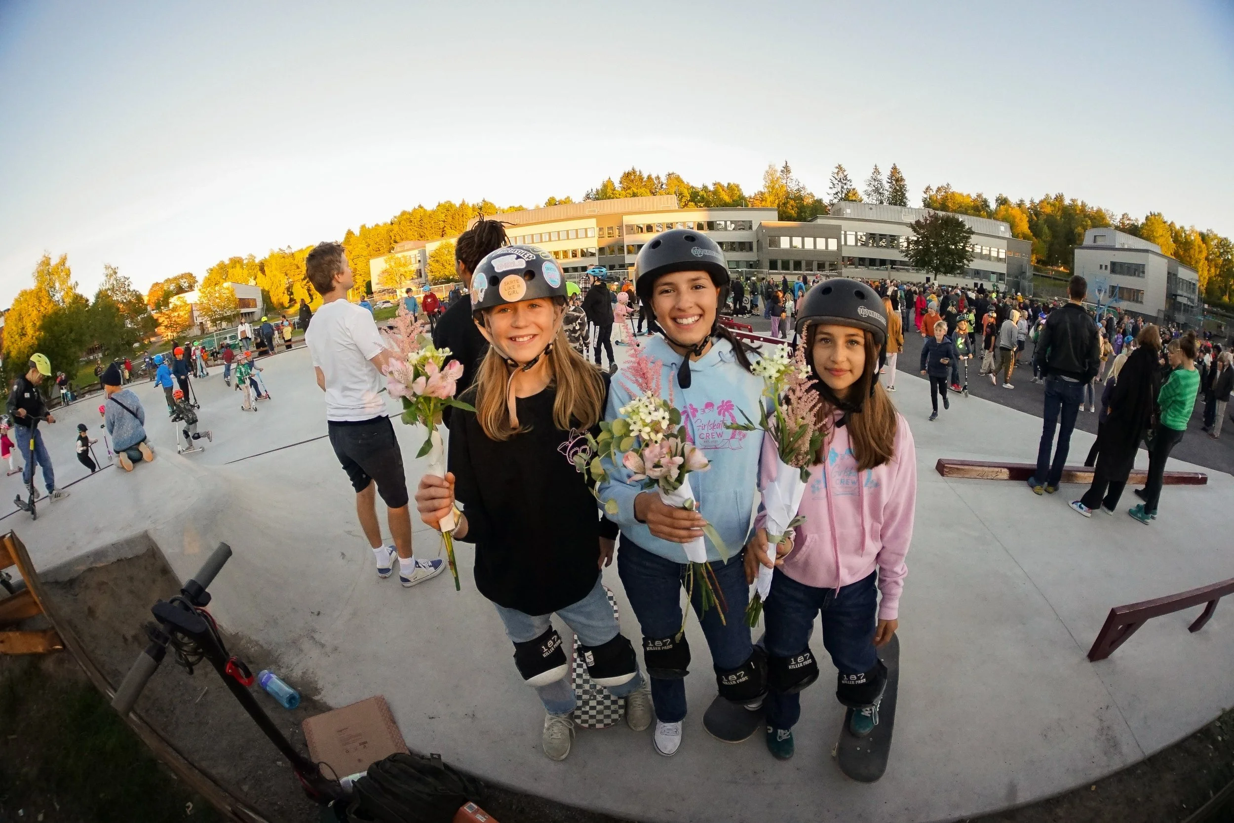 Three young girls wearing helmets holding bouquets of flowers at a skatepark during a sunny day, with many people in the background.