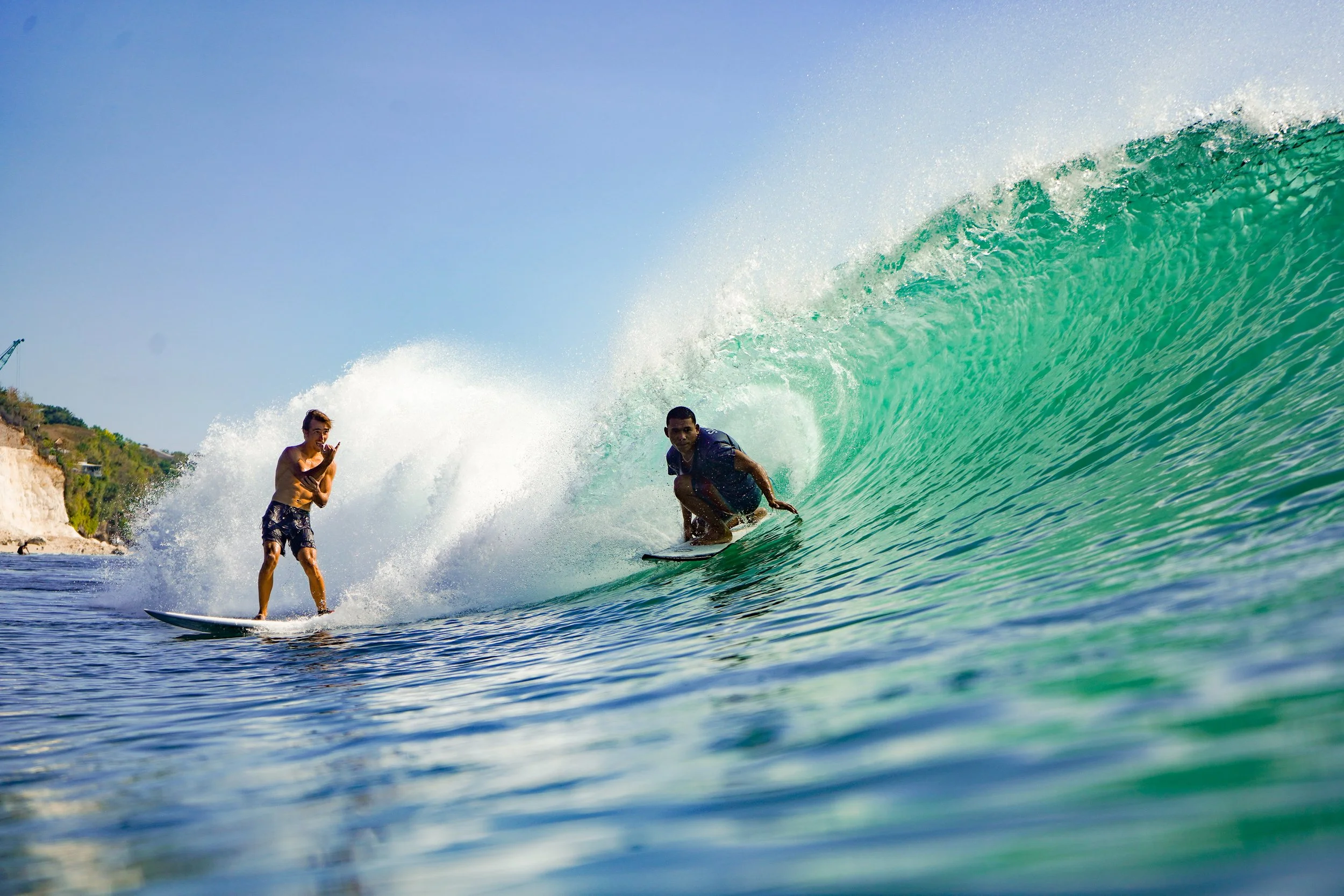 Two surfers riding a large wave in the ocean near a coastline with cliffs and greenery.