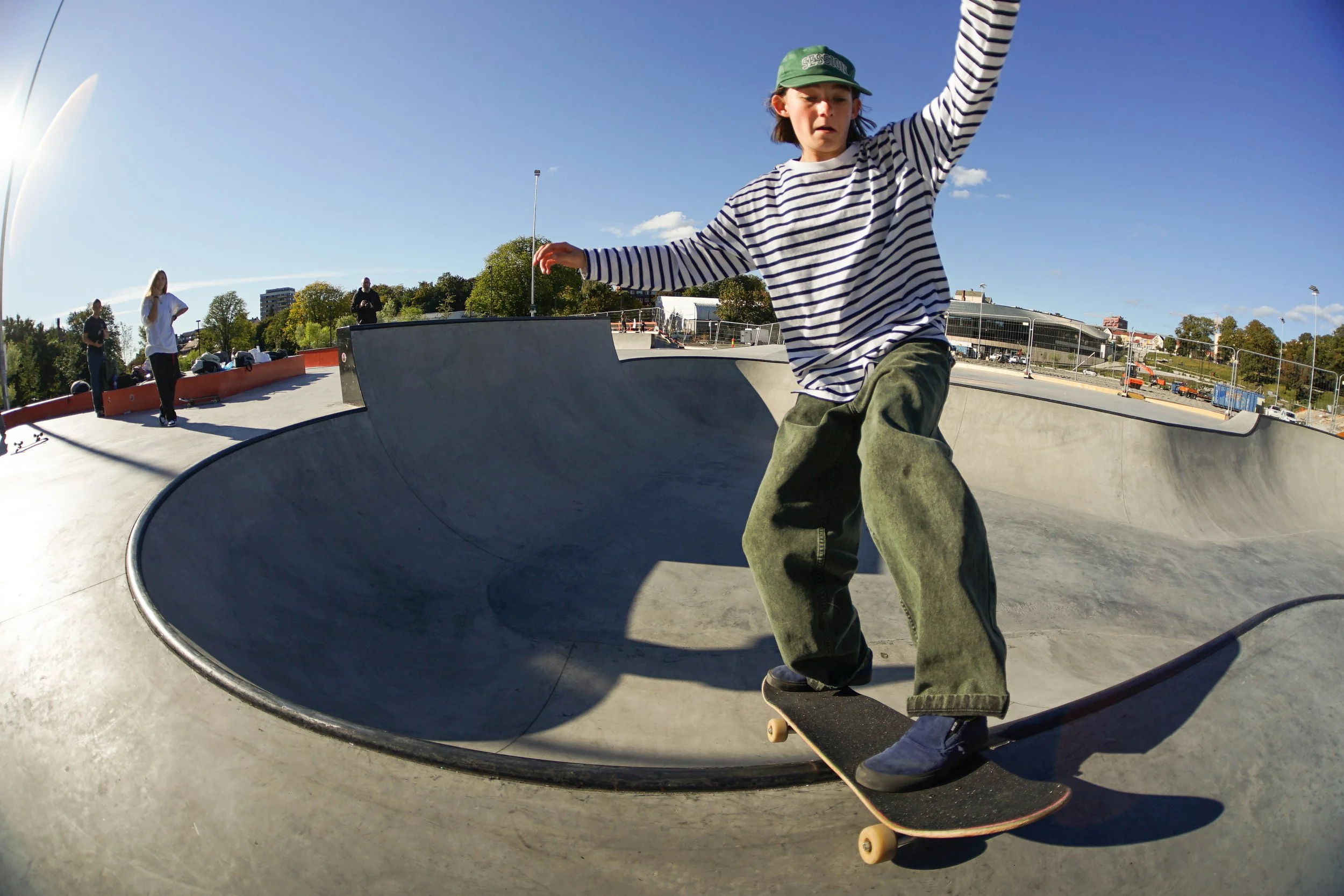 Young skateboarder in striped shirt and green pants performing a trick at an outdoor skate park on a sunny day.