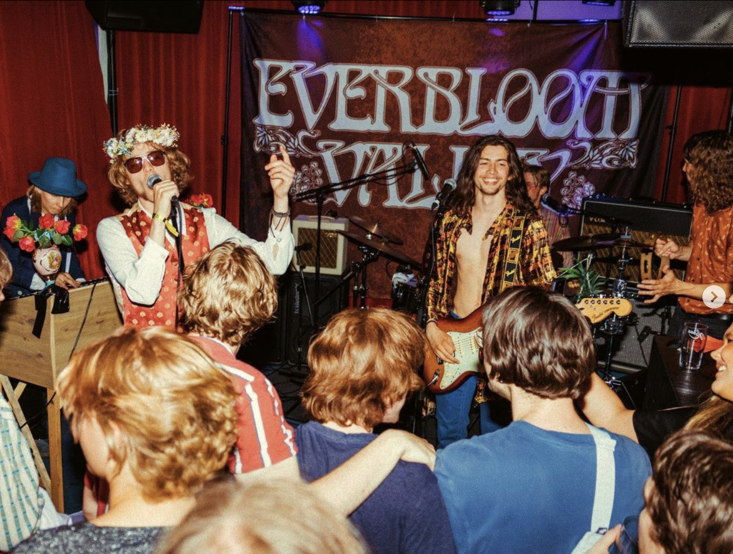 A band performing on stage with a backdrop reading "Everybody Live". The band members include a woman with a flower crown and sunglasses singing into a microphone, a man with long hair playing an electric guitar, and others playing instruments.