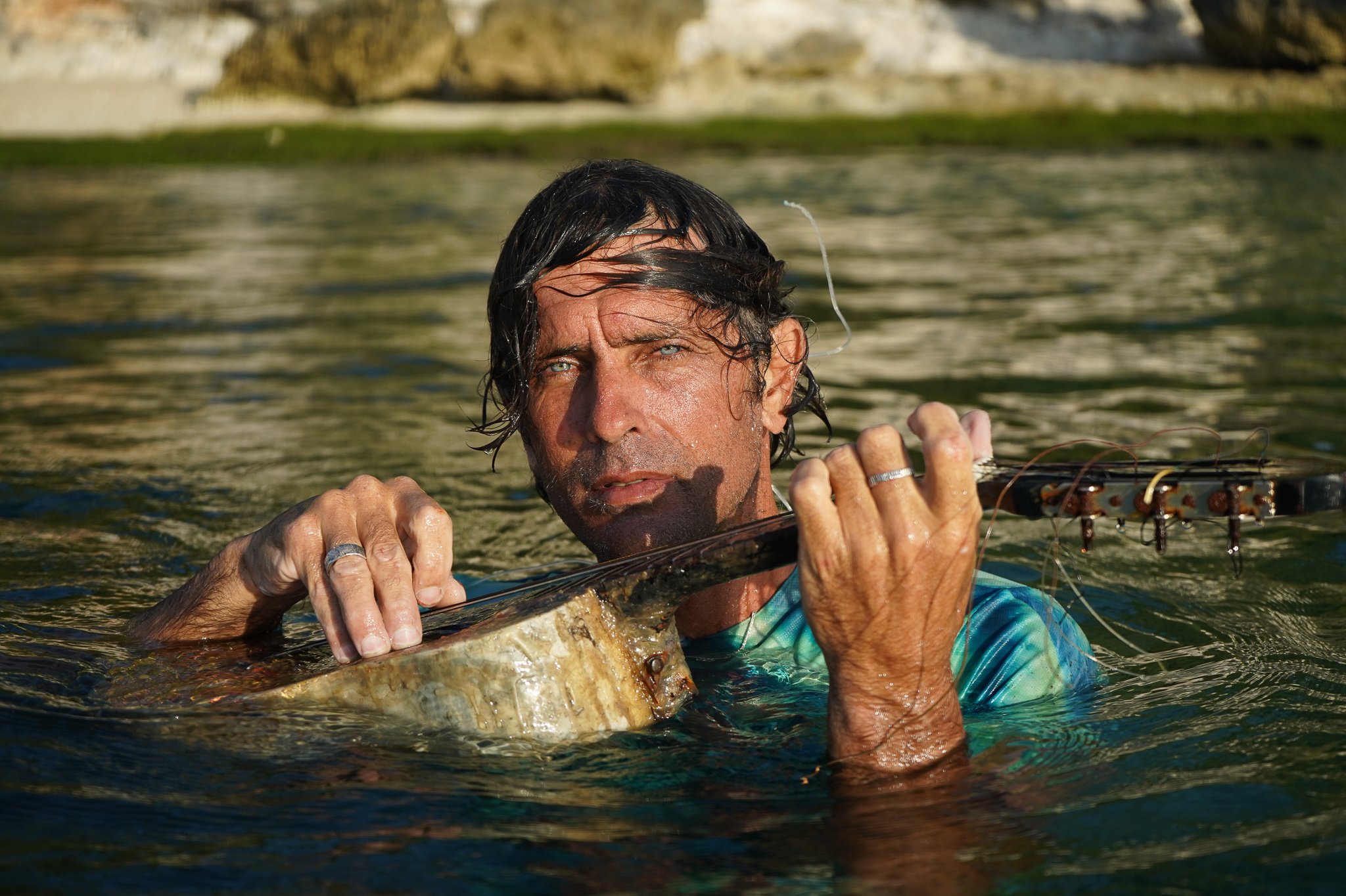 A man with wet hair and blue eyes holding a fishing rod while standing in water, with land and rocks in the background.