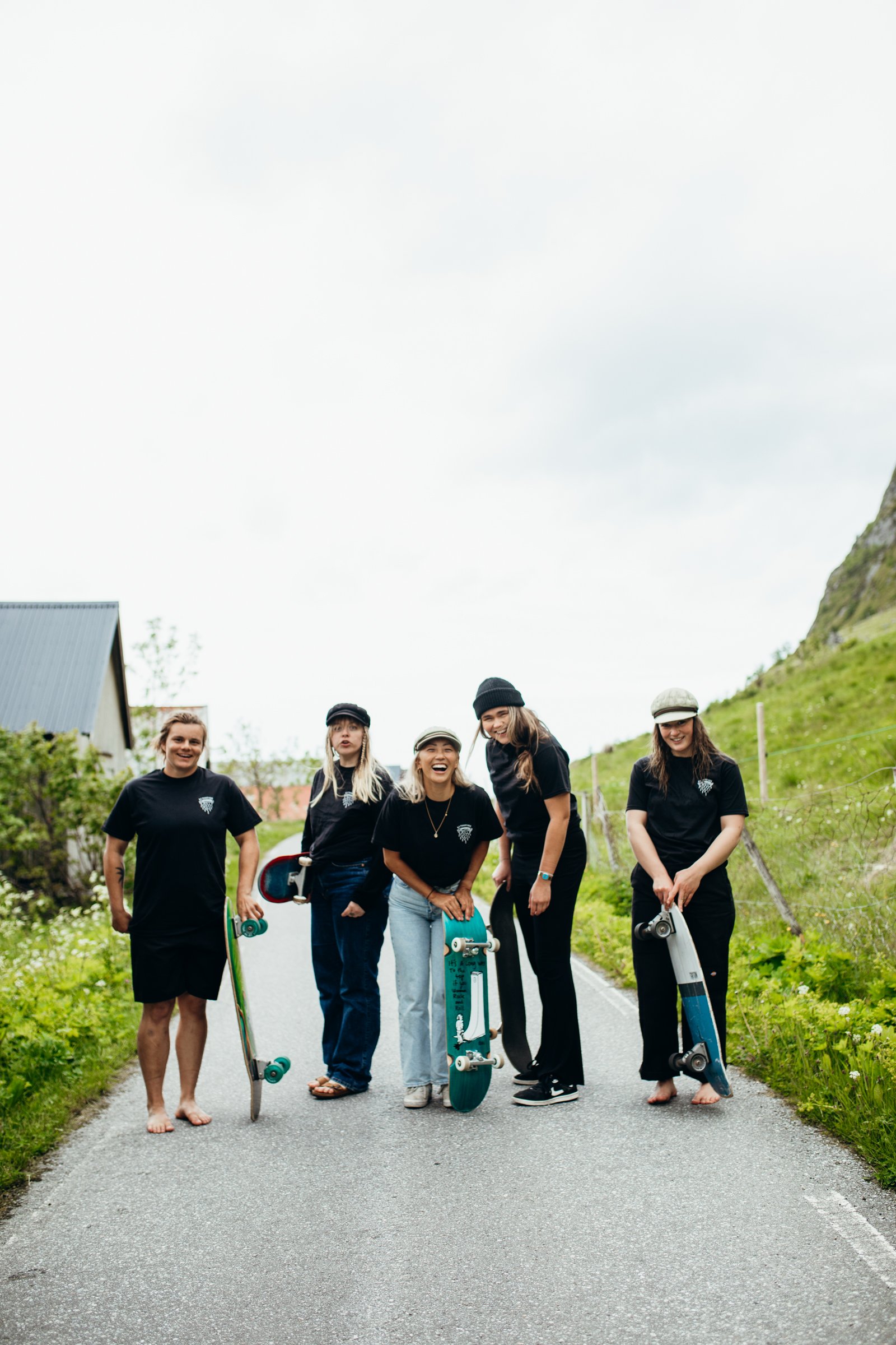 Five young women standing on a rural road, holding skateboards, smiling and enjoying a day outdoors.
