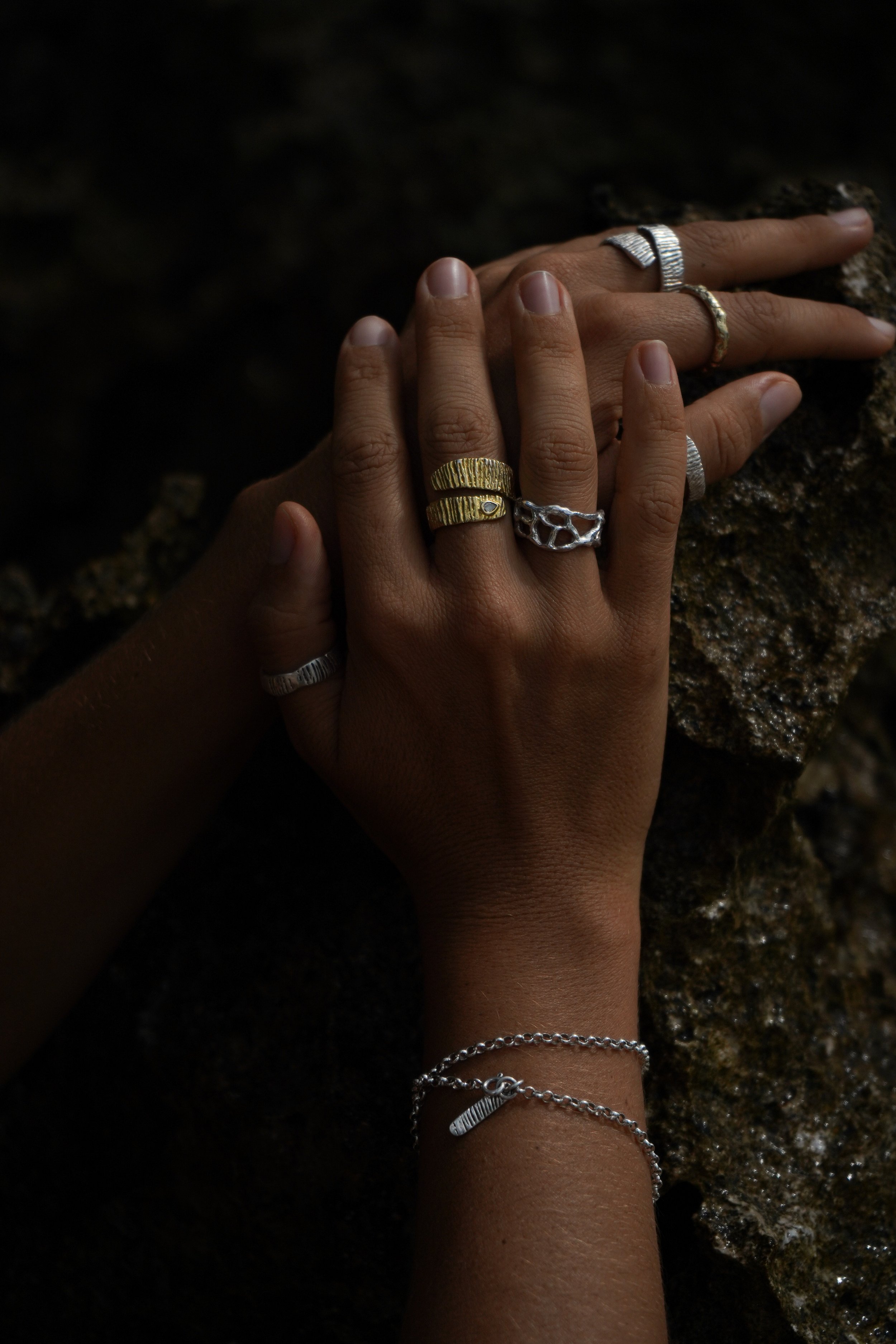 A person's hands and wrist decorated with multiple rings and a silver bracelet, resting on a textured dark stone surface.