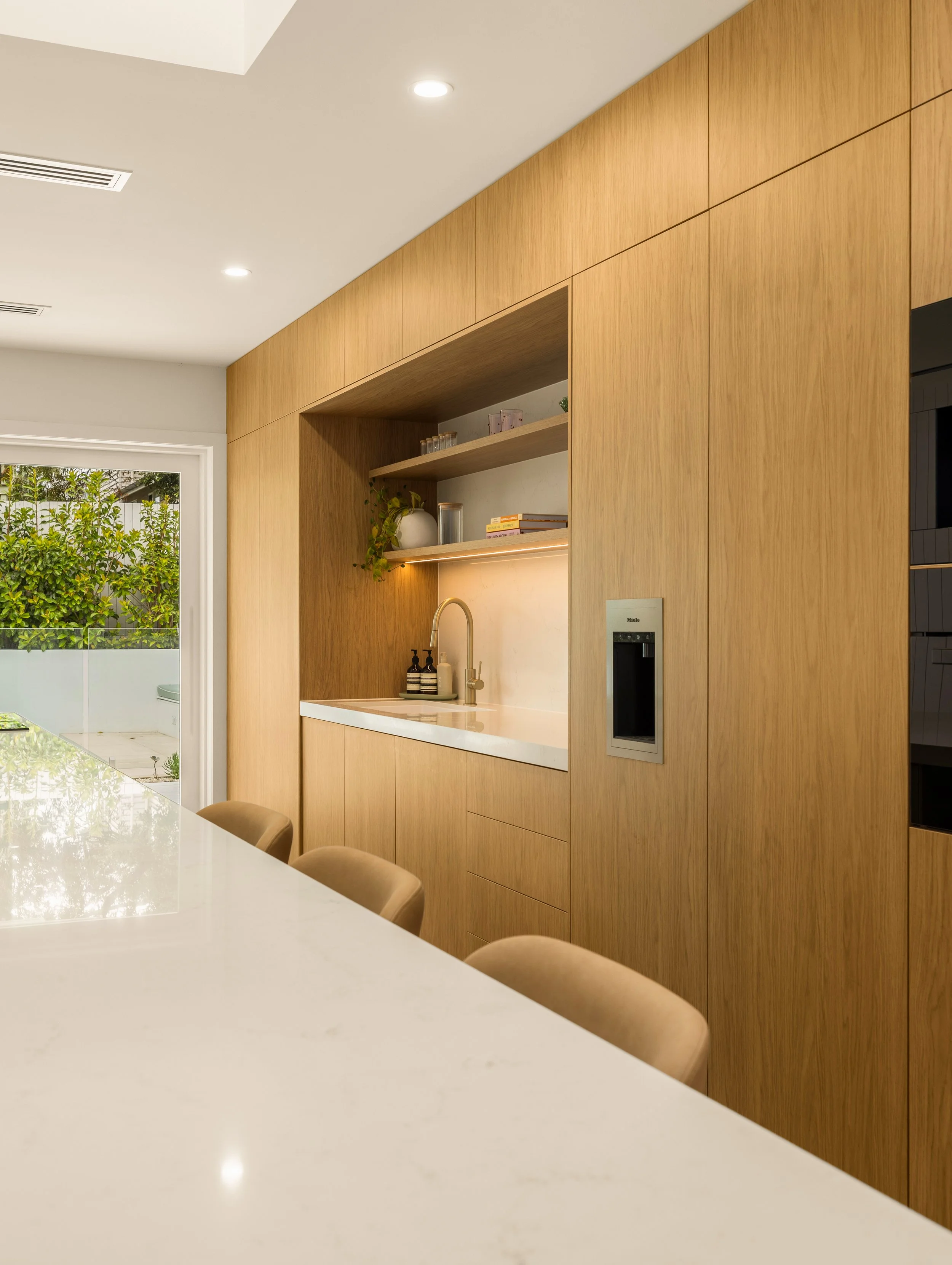 A modern kitchen with wood-paneled cabinets, a white countertop sink, a faucet, and a built-in water dispenser on the wall. There is a sliding glass door leading outside, and a white countertop island with tan chairs.
