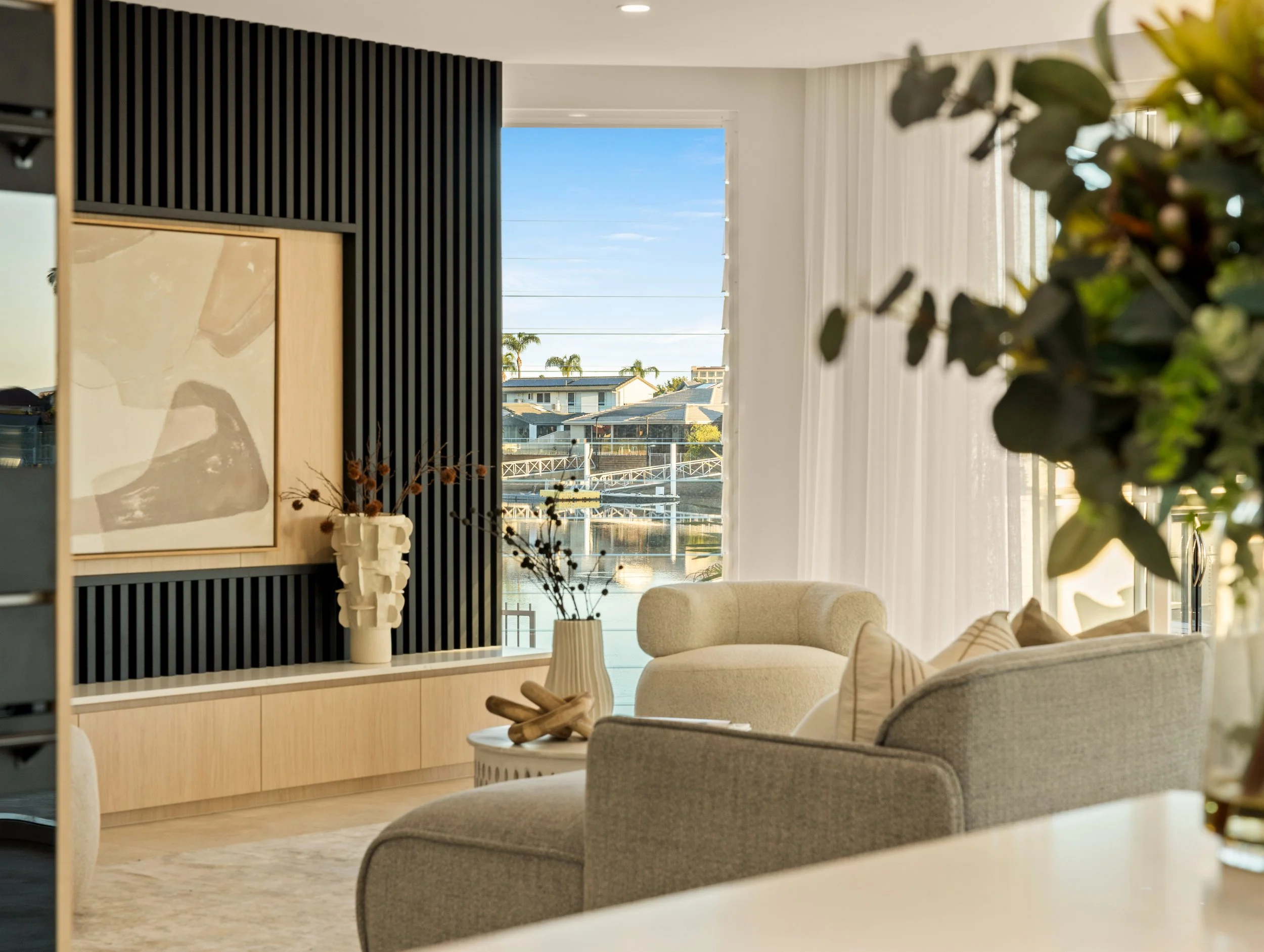 Living room with beige sofa, cream armchair, black slatted wall, framed artwork, vase with dried flowers, large window showing a waterway and houses outside, and white curtains.