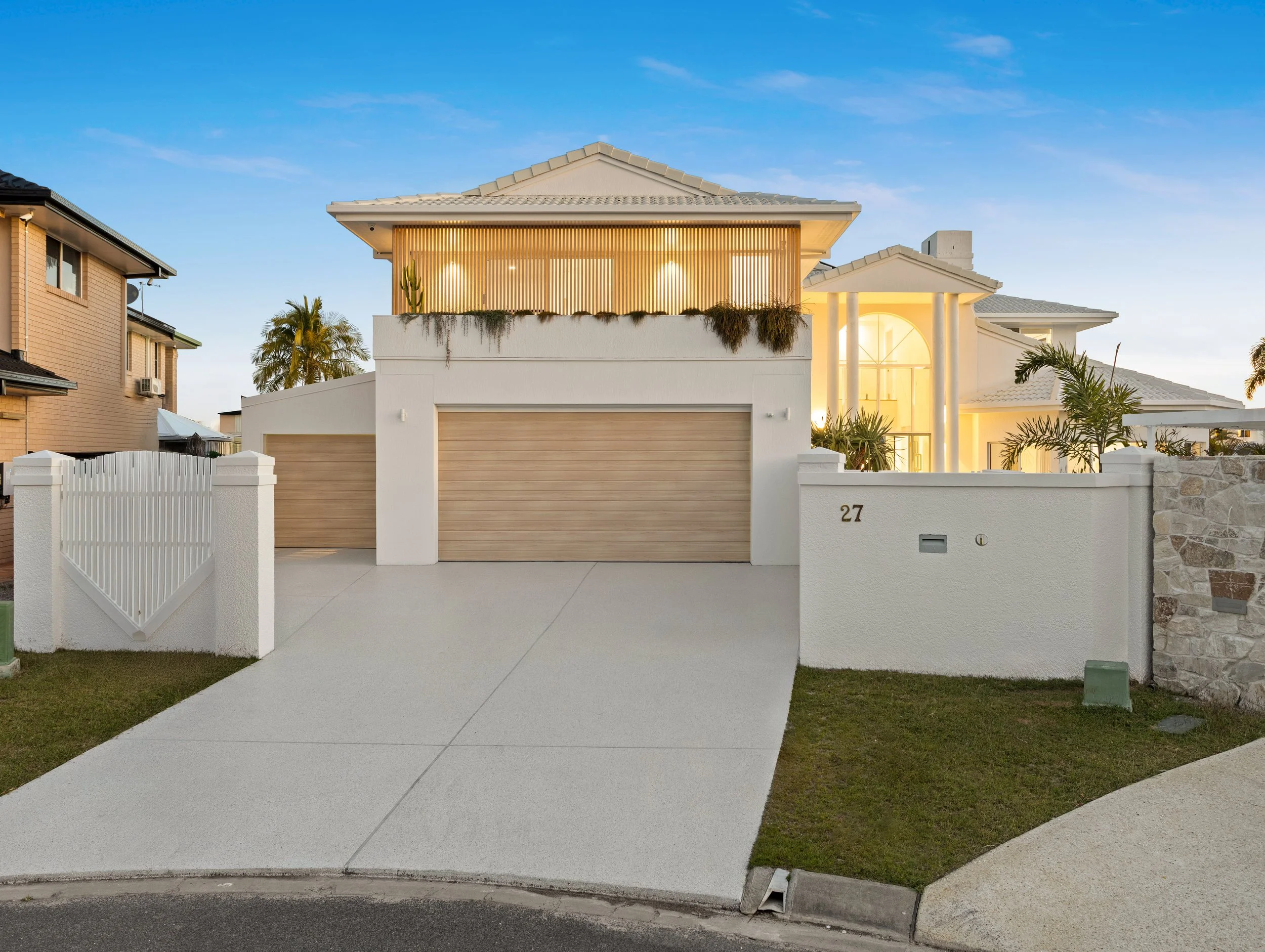 Modern residential house with white walls, wooden garage doors, a sloped roof, and tropical plants in the front yard, illuminated by warm lighting at dusk.
