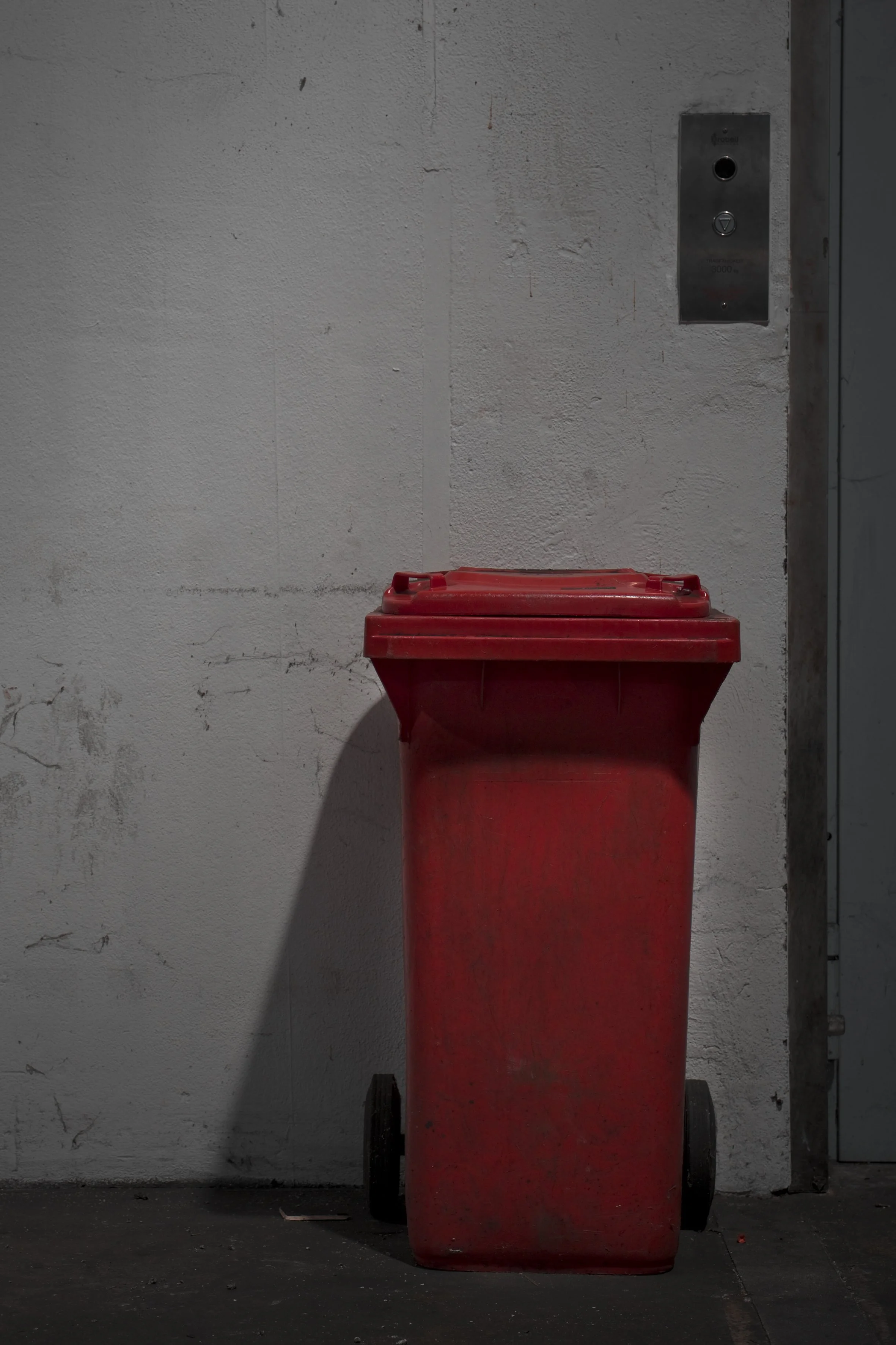 Roter Abfallcontainer mit schwarzen Rädern vor einer weißen Wand neben einem Aufzugsschalter.
