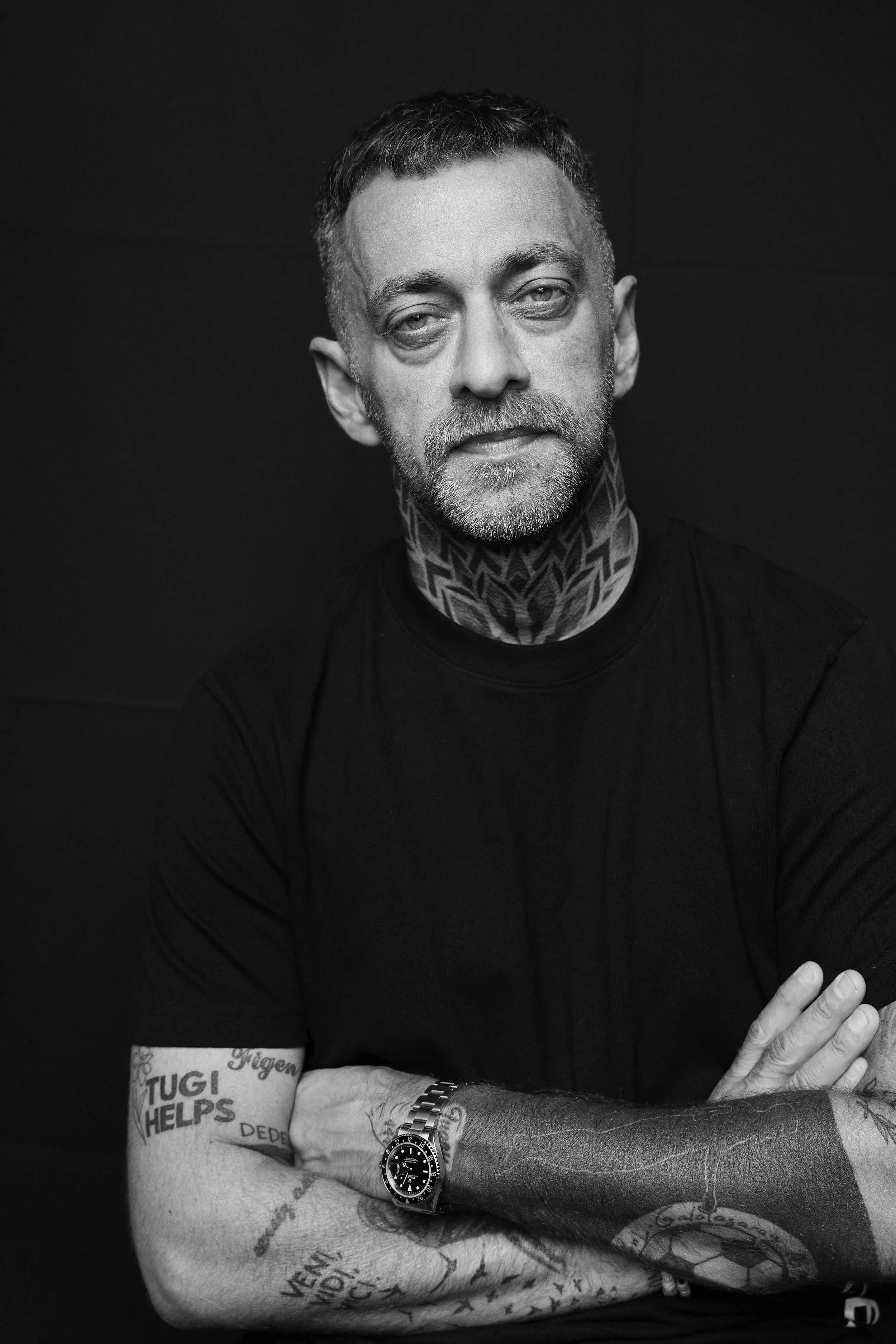 Schwarz-weiß-Porträt eines Mannes mit Tattoos, Beard und Uhren, der die Arme verschränkt trägt ein schwarzes T-Shirt gegen einen dunklen Hintergrund.