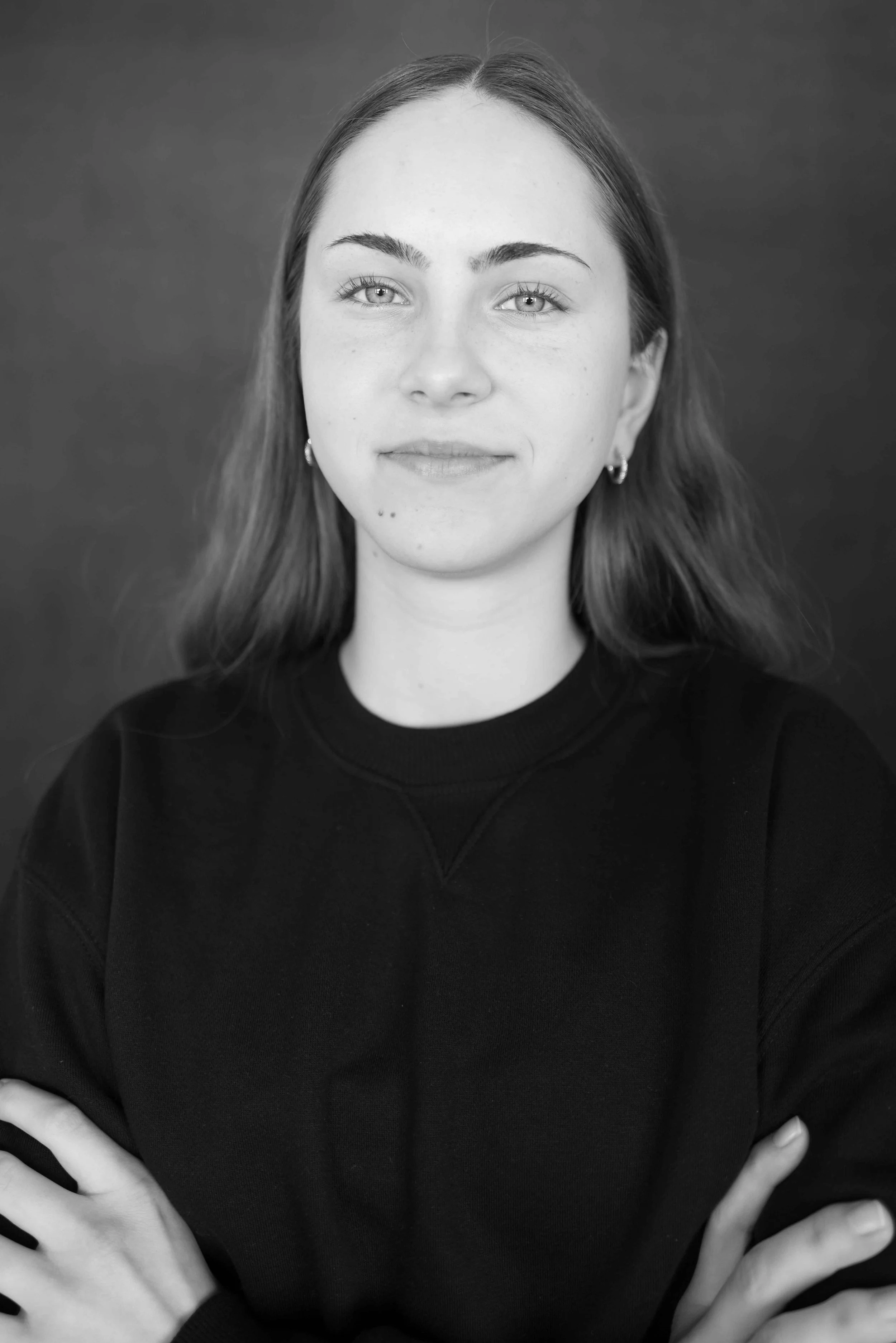 Schwarz-Weiß-Fotografie einer jungen Frau mit gerader, schulterlanger Haar, getragen in einem dunklen Pullover, die die Arme verschränkt und direkt in die Kamera blickt.