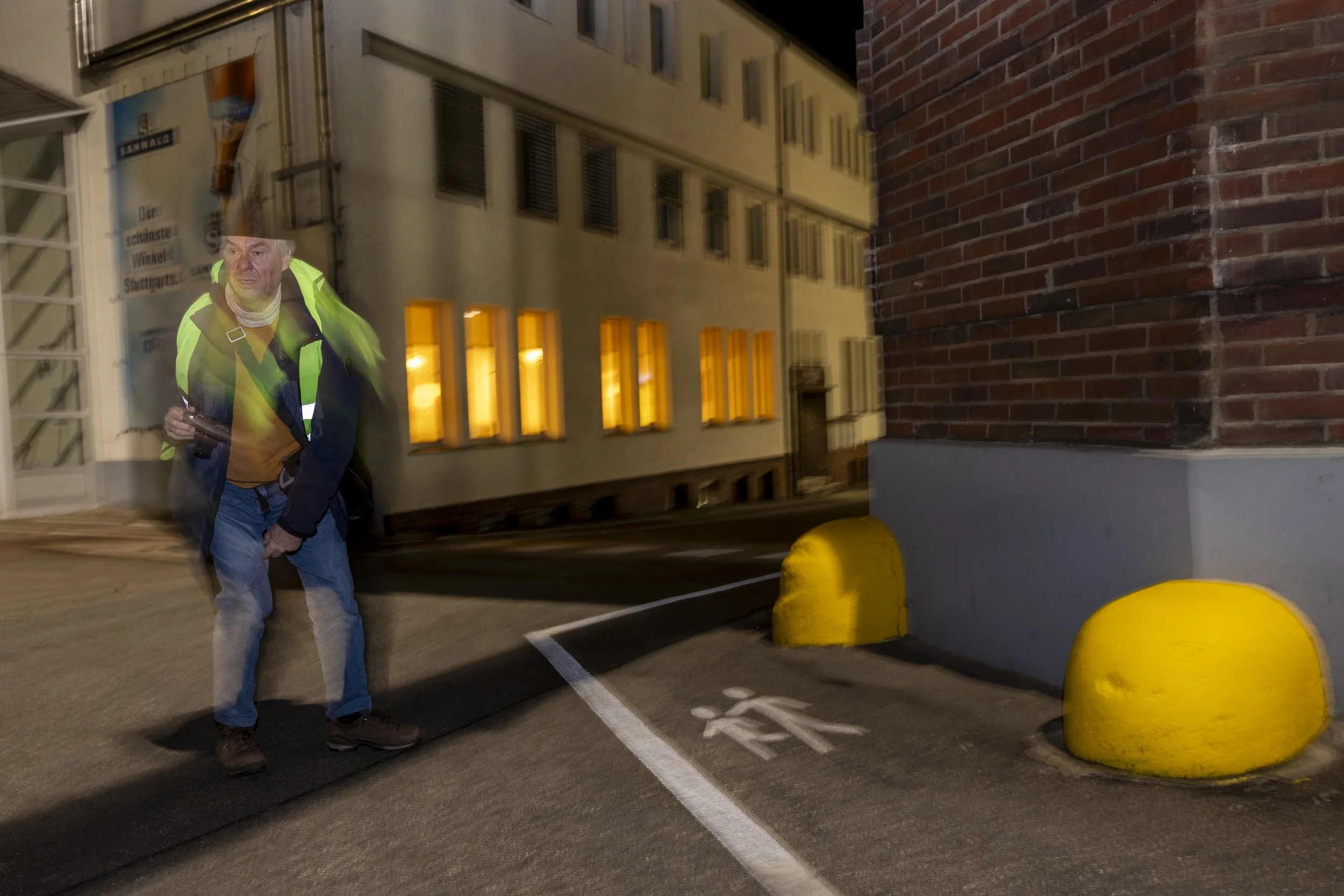 Nachtaufnahme eines Mannes in Warnweste, der im Kreis läuft, vor einem Gebäude mit leuchtenden Fenstern. Er ist leicht verschwommen, als ob er sich bewegt.