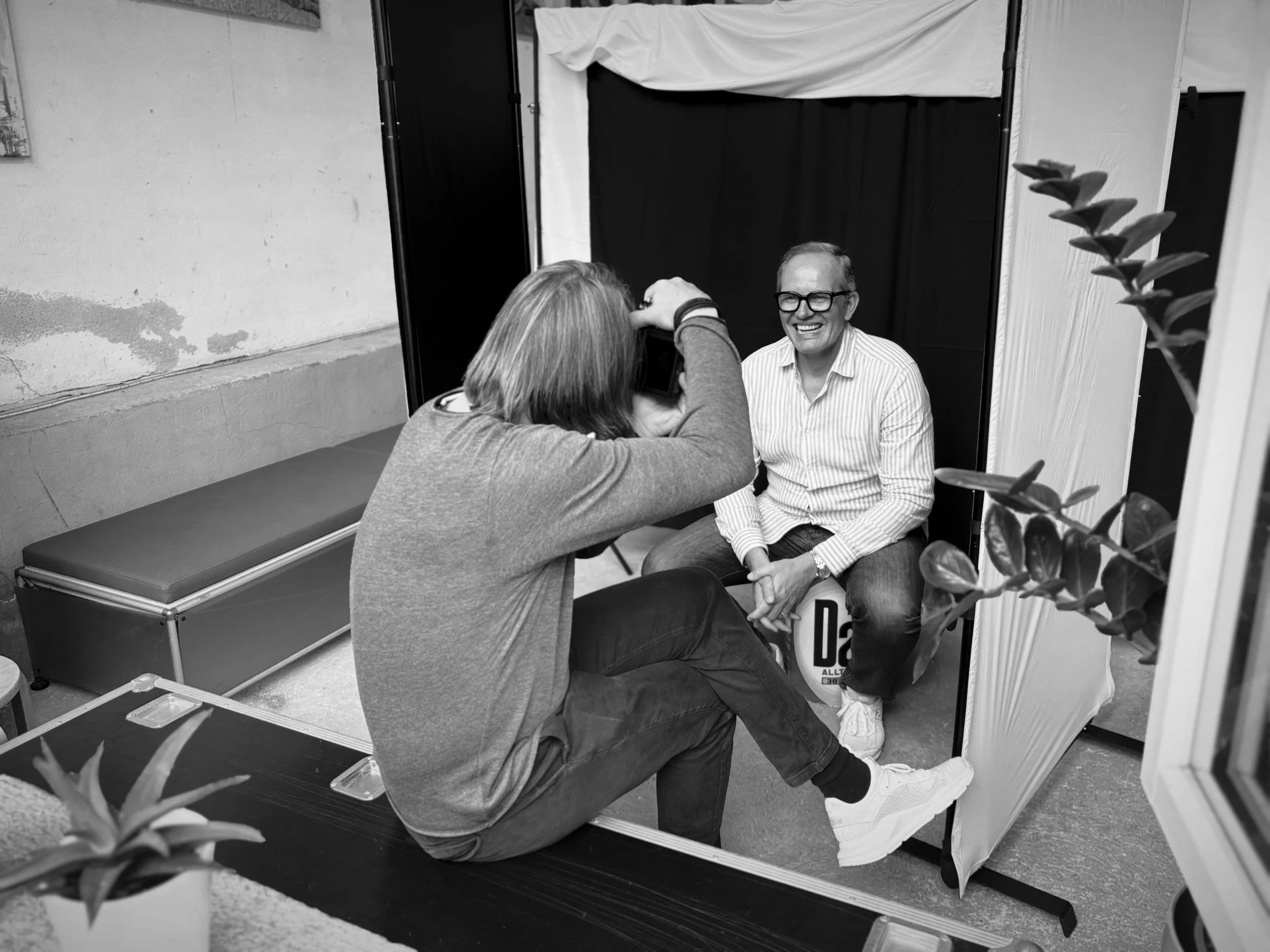 Ein Mann mit Brille sitzt vor einem schwarzen Hintergrund, während eine Person mit Schulterlänge Haaren ein Foto von ihm macht.