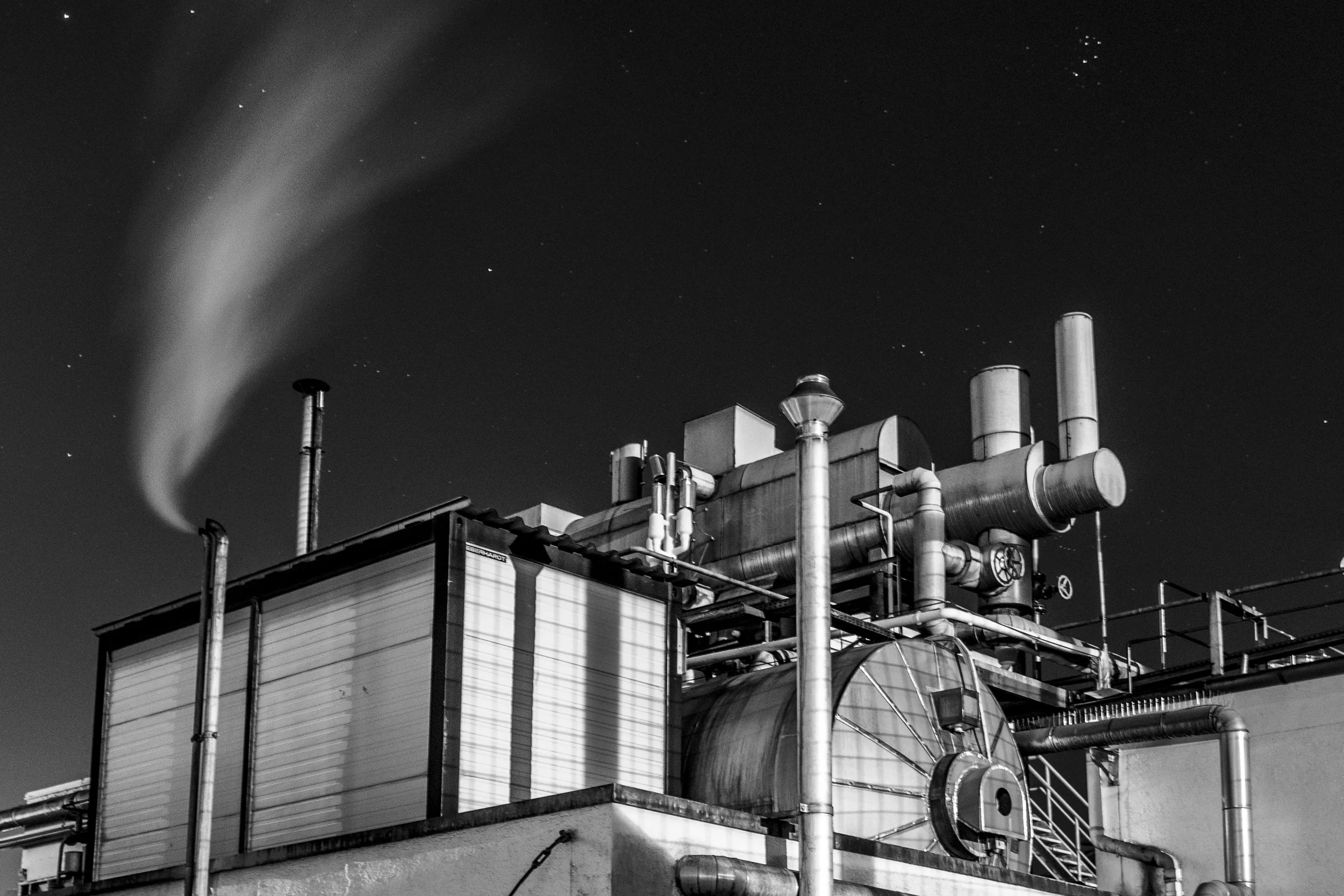 Schwarz-weiß-Foto eines Industriegebäudes mit Rohrleitungen und Schornstein, aus dem Rauch aufsteigt, bei Nacht.