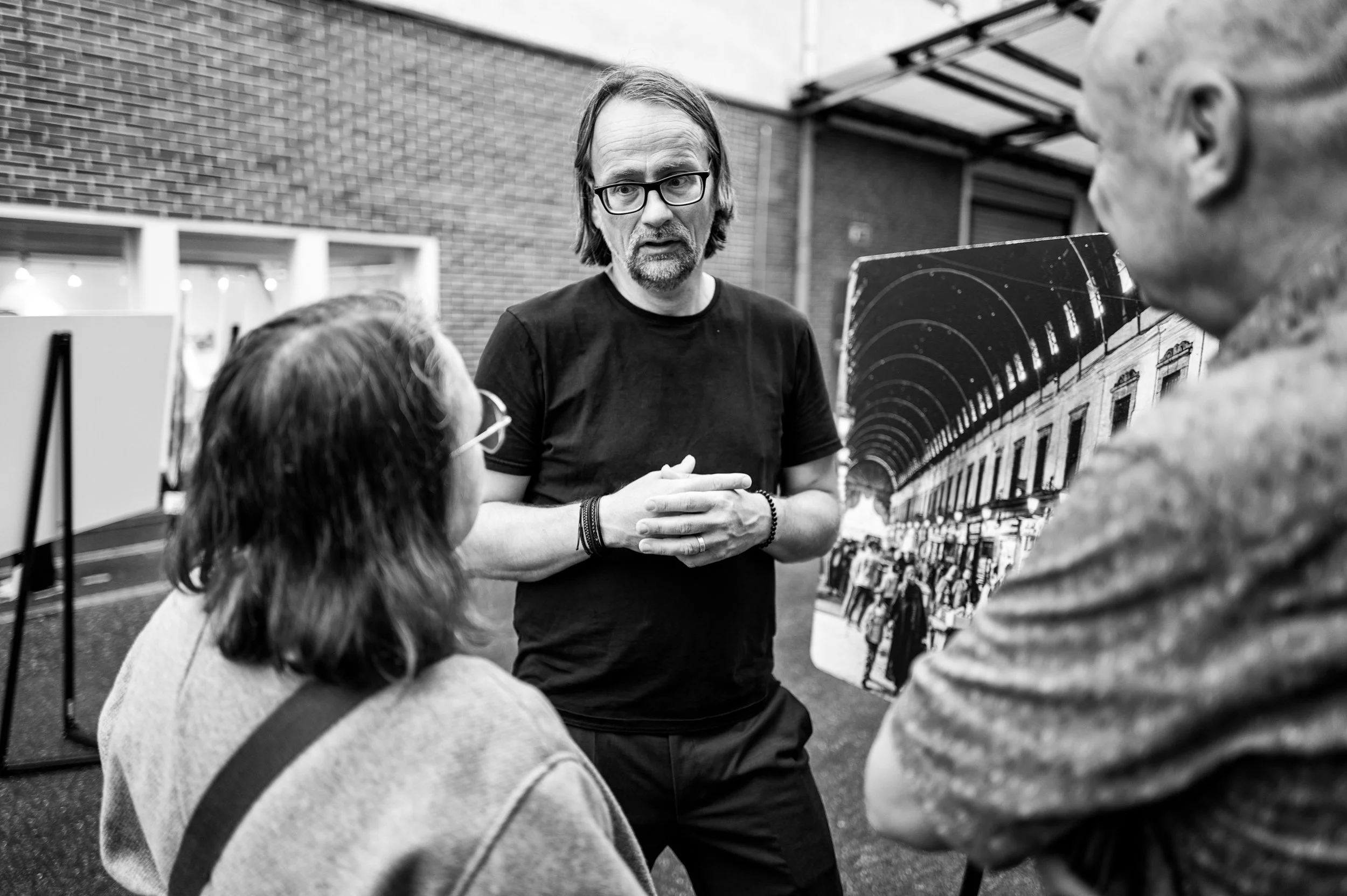 Drei Personen im Gespräch, eine Person erklärt an einem Display eine historische Fotografie in einem Kunst- oder Ausstellungsraum.