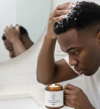 A young man applying hair product to his hair while looking in a mirror, with soap lather on his head, in a bathroom.