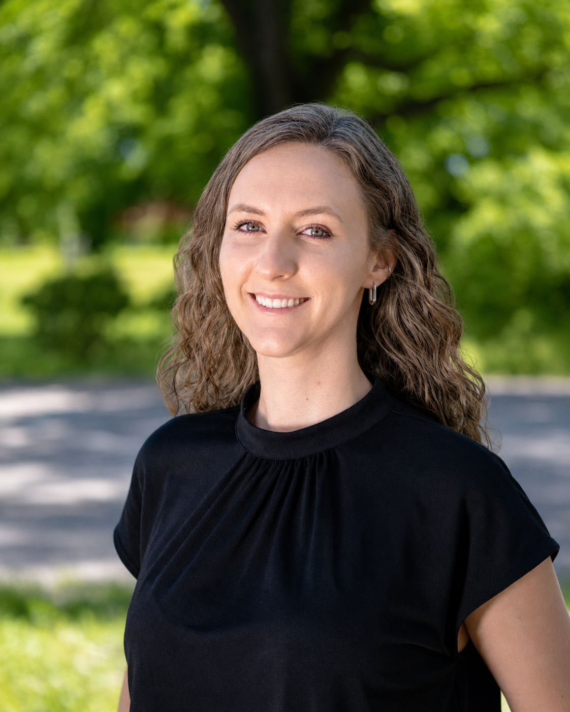 Portrait of a smiling woman with curly hair, wearing a black top, standing outdoors with green trees in the background.